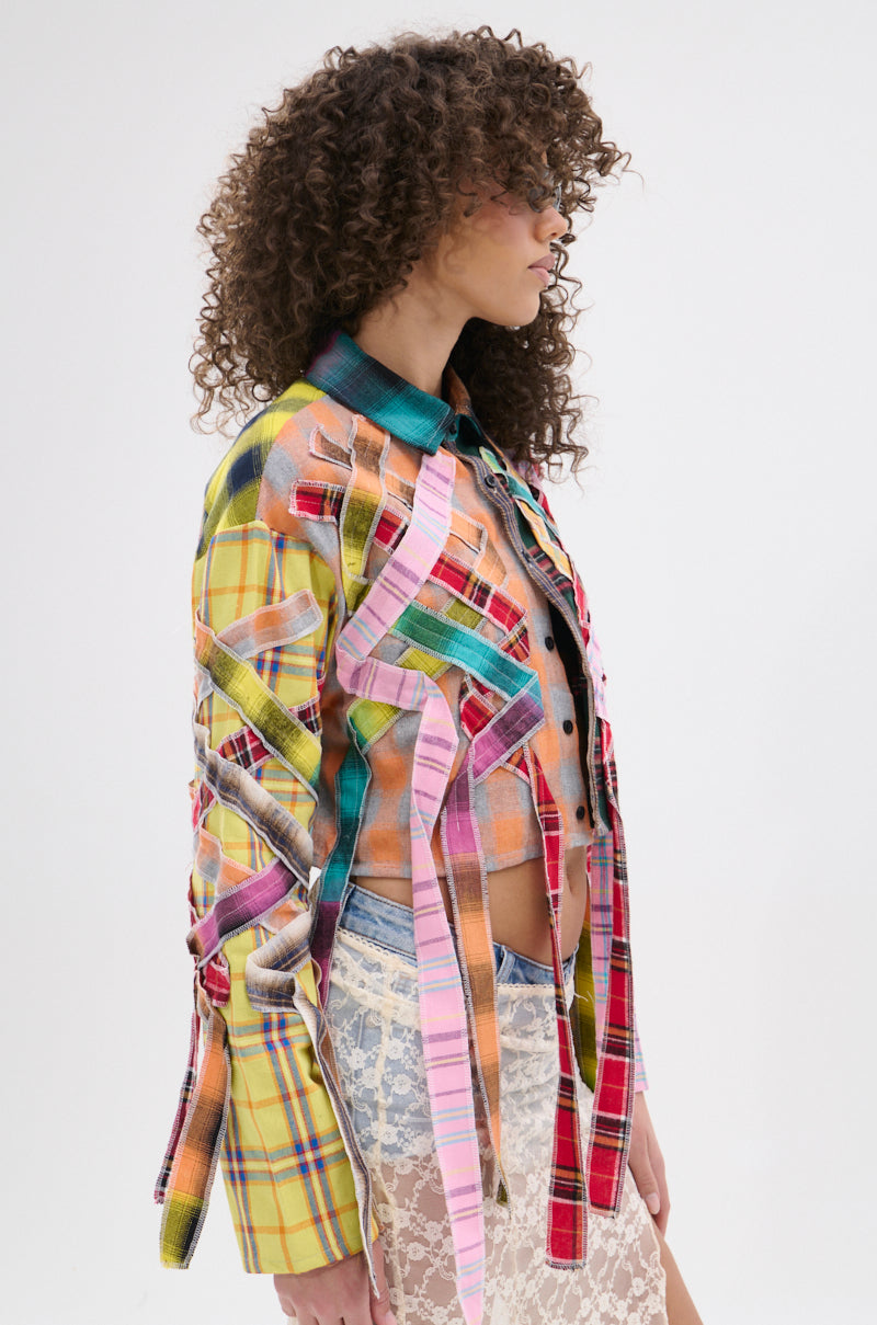 A woman with curly hair stands in profile against a white background, wearing the FEELING CRAZY MIXED PLAID BUTTON DOWN—a punk-inspired, colorful patchwork plaid jacket with pink ribbon accents—paired with a white lace skirt.