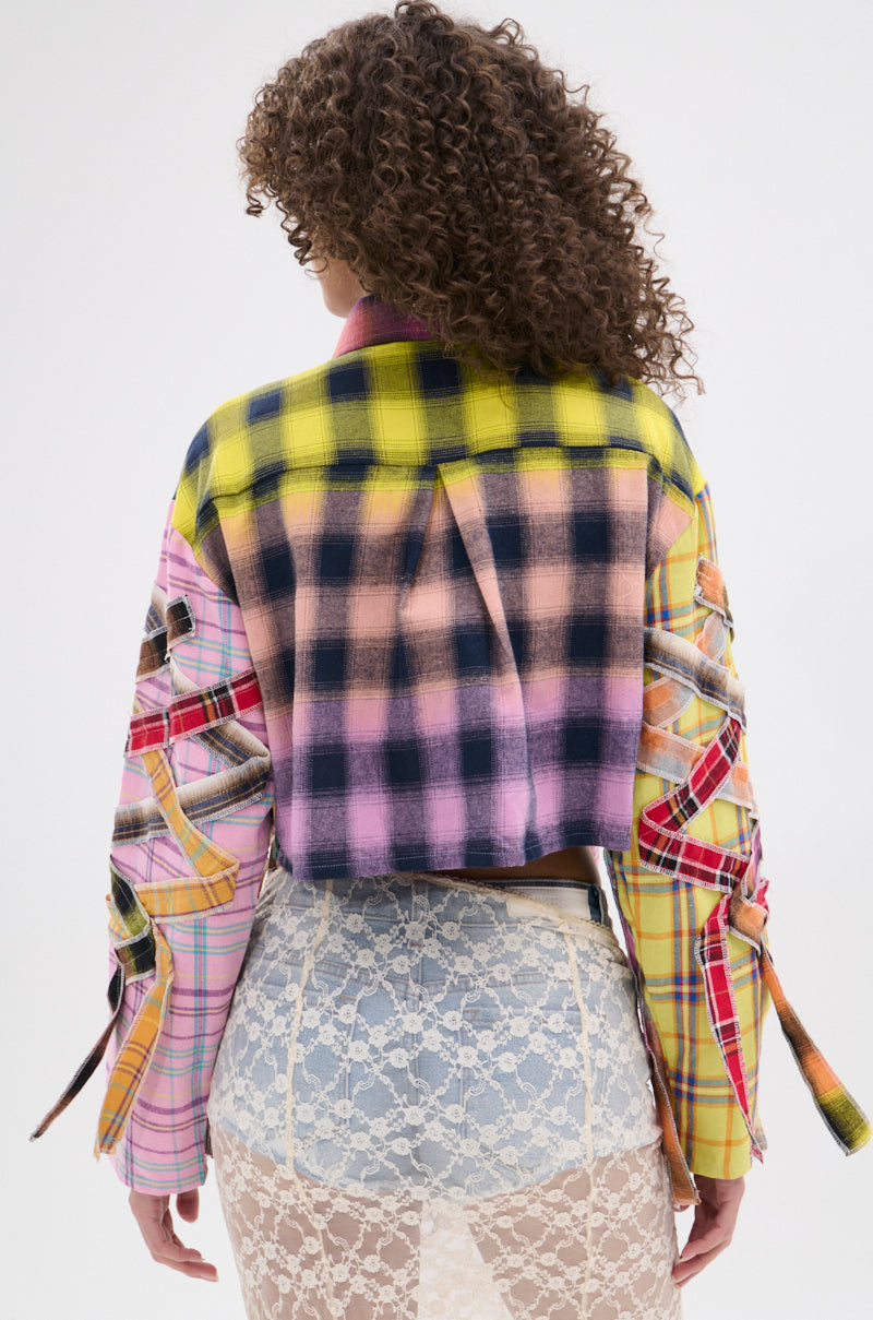 A person with curly hair, seen from behind, wears the FEELING CRAZY MIXED PLAID BUTTON DOWN, styled in a punk-inspired way with cut-out details on the sleeves and back, paired with a sheer white lace skirt over light blue shorts.