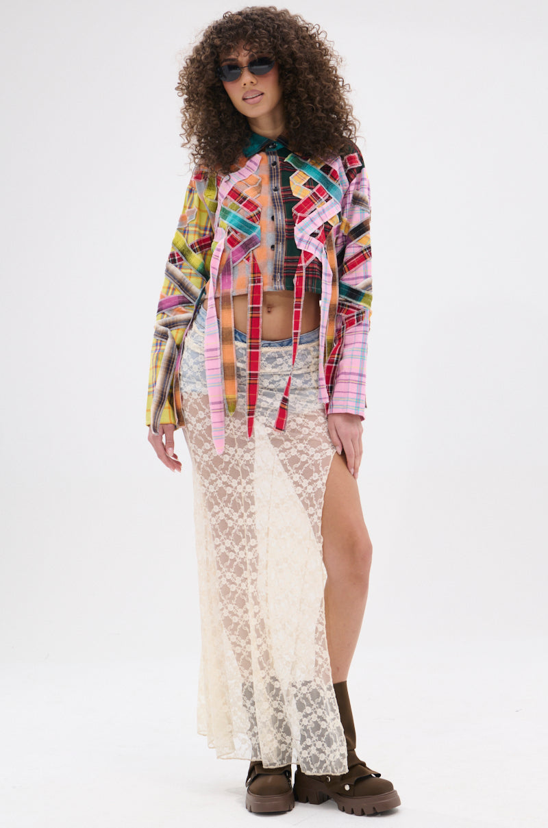 A woman with curly hair and sunglasses wears the FEELING CRAZY MIXED PLAID BUTTON DOWN over a plaid bralette, styled with a long sheer white lace skirt and chunky brown boots, standing against a white background.