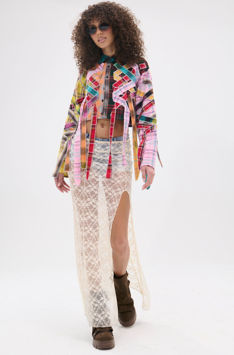 A person with curly hair and sunglasses wears the FEELING CRAZY MIXED PLAID BUTTON DOWN, a sheer white lace skirt with a thigh-high slit, and brown boots, confidently walking against a white background.