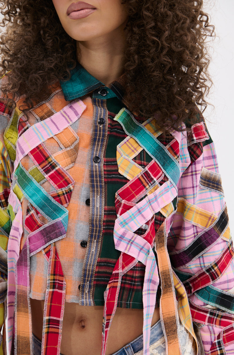 A woman with curly hair wears the FEELING CRAZY MIXED PLAID BUTTON DOWN, a cropped, punk-inspired shirt with colorful patchwork plaid and frayed, hanging fabric strips. Only her head and torso are visible.