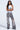 A woman with long hair and glasses stands with arms crossed, wearing a white tank top, blue-patterned design, and LETS GET IT GOING DISTRESSED JOGGER in gray. Her edgy streetwear style is perfectly complemented by the light neutral background.