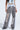 A person is sporting the LETS GET IT GOING DISTRESSED JOGGER in gray with rips and frayed hems, paired with a light blue cropped top featuring a metallic buckle design and black chunky shoes. The scene is set against a plain, well-lit background, creating a standout streetwear look.