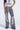 A person in a sleeveless top with corset detail and LETS GET IT GOING DISTRESSED JOGGER jeans, featuring white patches and heavy rips at the knees and thighs, stands in black platform shoes. Rings adorn the hand, creating a bold streetwear statement.