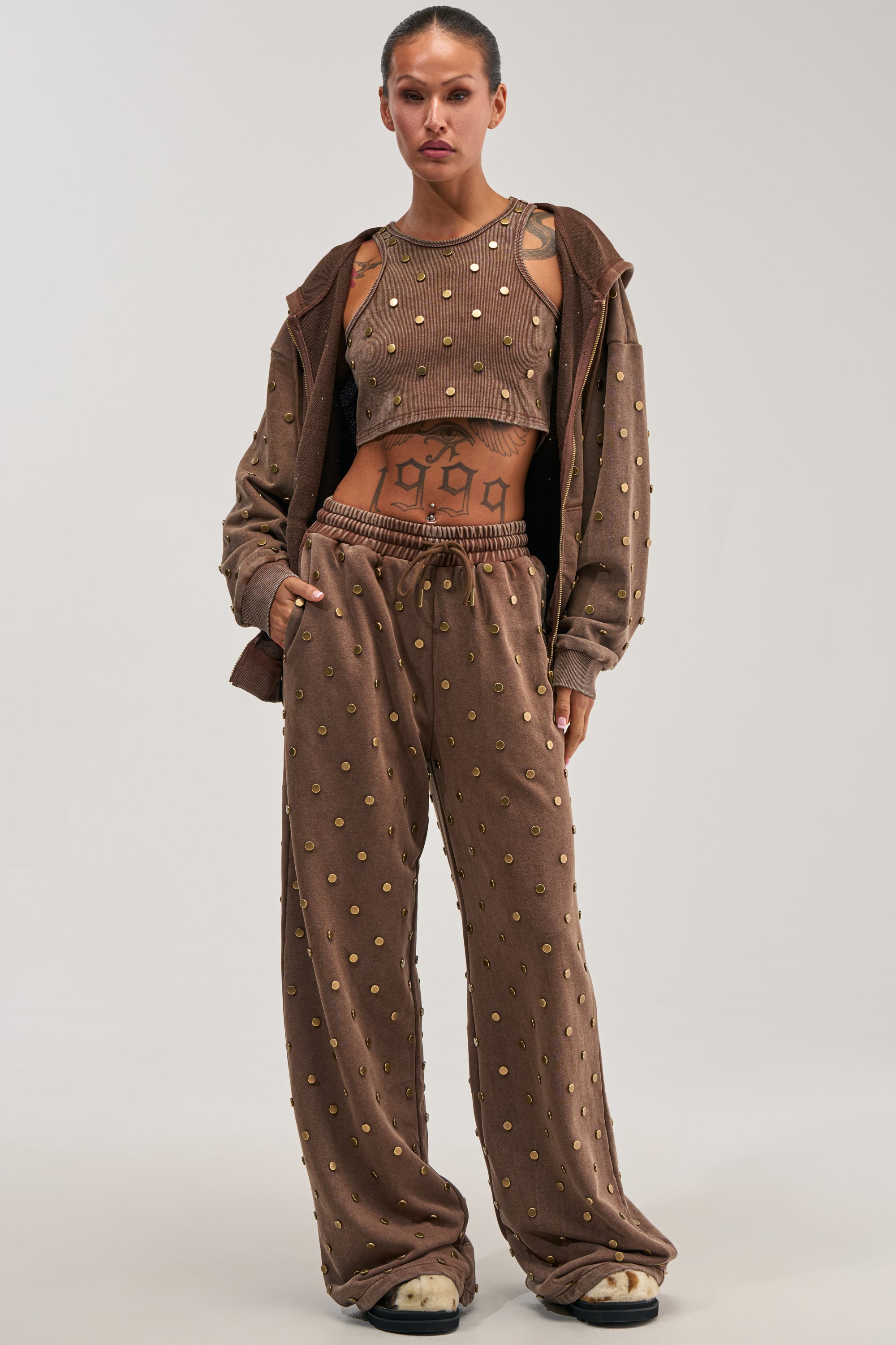 Model showcases the POPPING OFF WIDE LEG JOGGER IN BROWN, styled with a matching polka dot crop top and jacket. The loose-fitting set highlights visible tattoos on the midriff, exuding undeniable confidence and an effortlessly cool streetwear vibe against a plain backdrop.