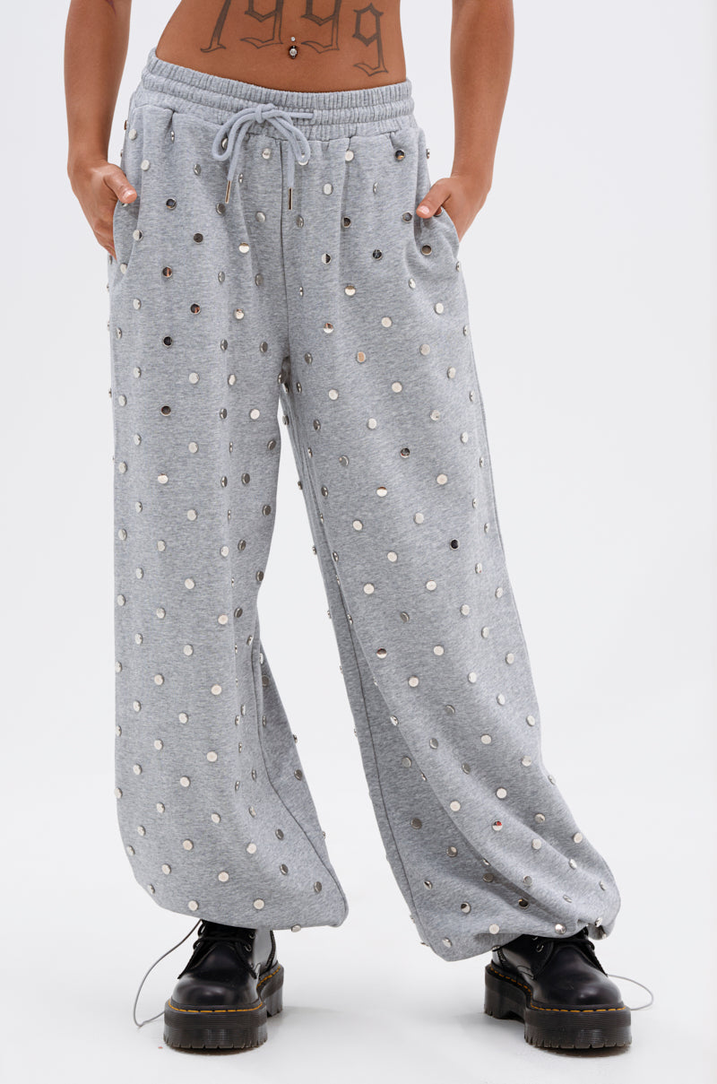 A person wears the POPPING OFF WIDE LEG JOGGER—loose-fitting, gray, high-rise joggers with silver studs—paired with black chunky lace-up shoes and a visible torso tattoo. Their hands are in their pockets against a white background. 1
