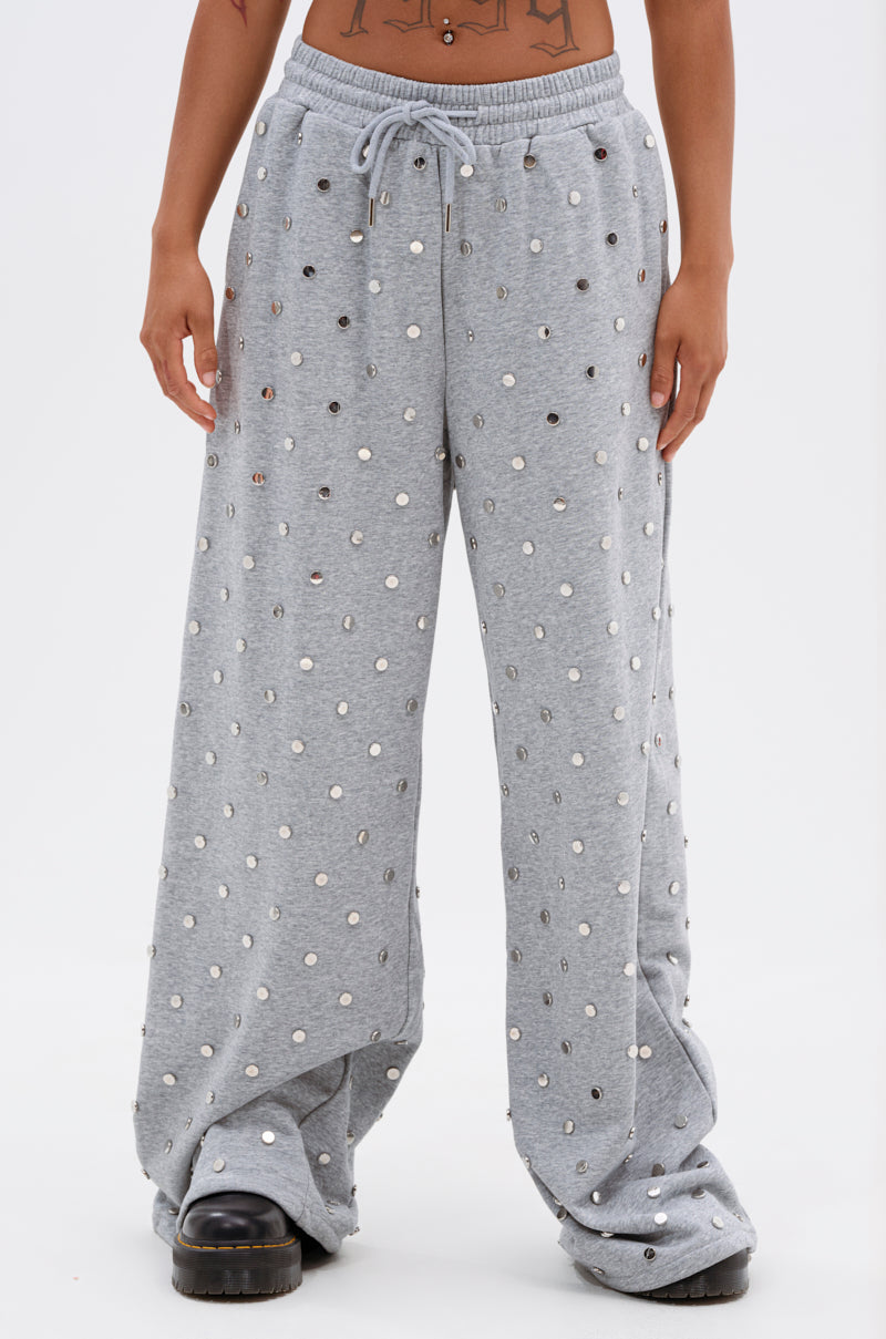 Wearing the POPPING OFF WIDE LEG JOGGER, a loose, light gray high-rise sweatpant with silver sequins and an elastic drawstring waistband, paired with black platform shoes. The person's midriff and tattoos are visible. 2
