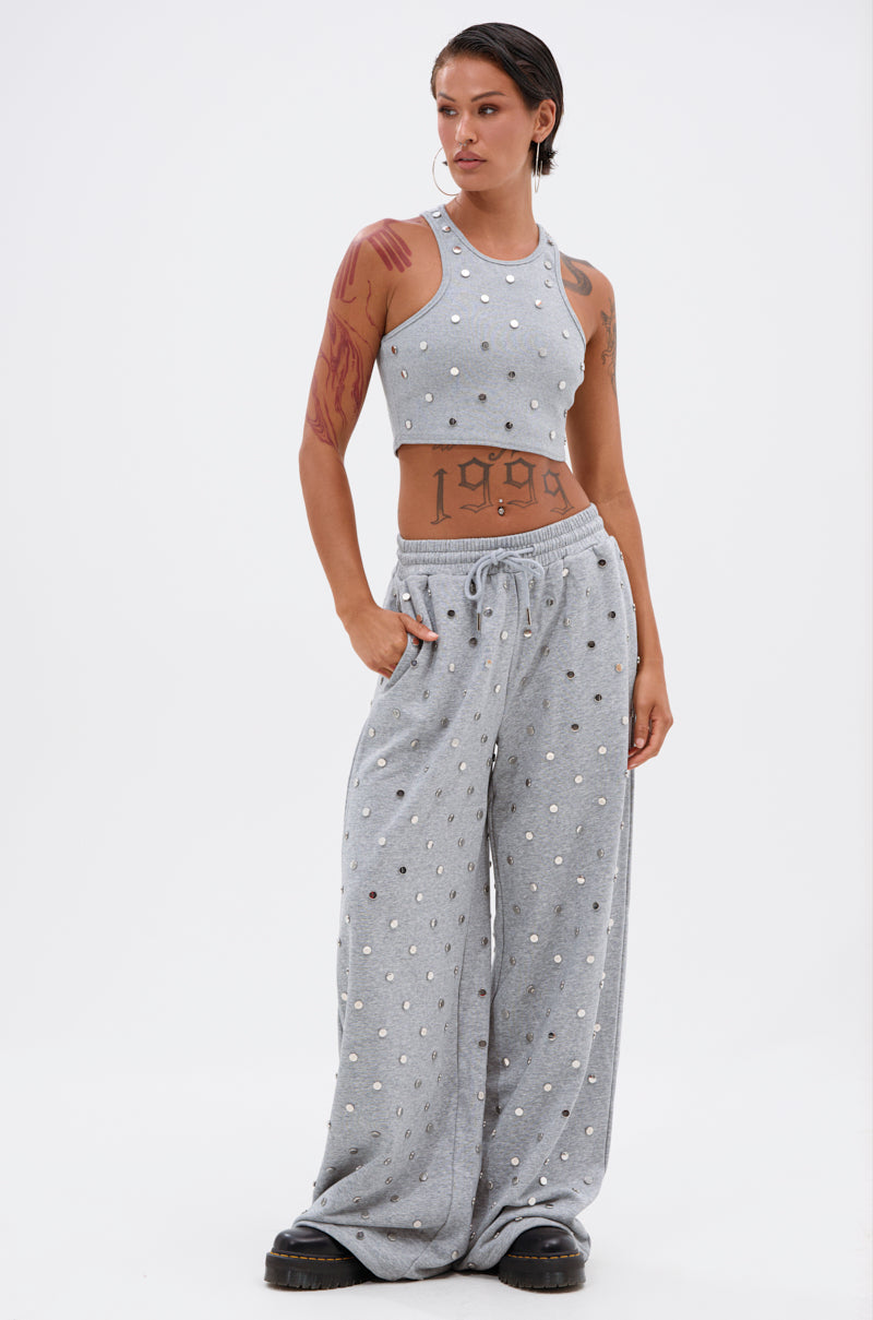 A person with short hair and visible tattoos poses confidently in the POPPING OFF WIDE LEG JOGGER—grey, wide-leg pants adorned with white beads and studs—paired with a matching sleeveless crop top and black chunky shoes against a white backdrop. 6