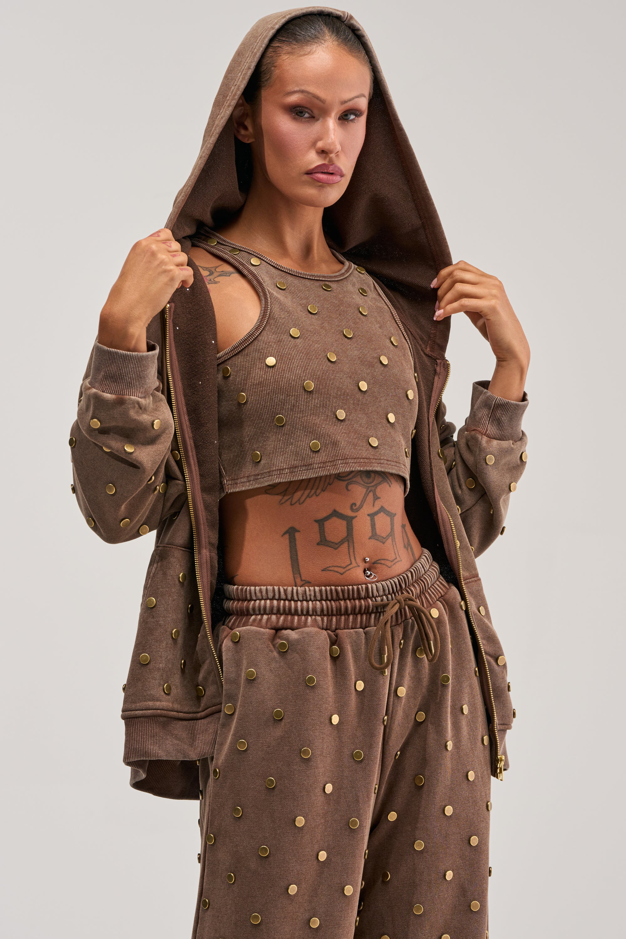 A woman models the POPPING OFF ZIP UP SWEATSHIRT IN BROWN, featuring gold stud detailing. She pairs it with a crop top and sweatpants for an effortlessly cool streetwear loungewear look, holding the hood with both hands to reveal her torso tattoos against a plain light background.