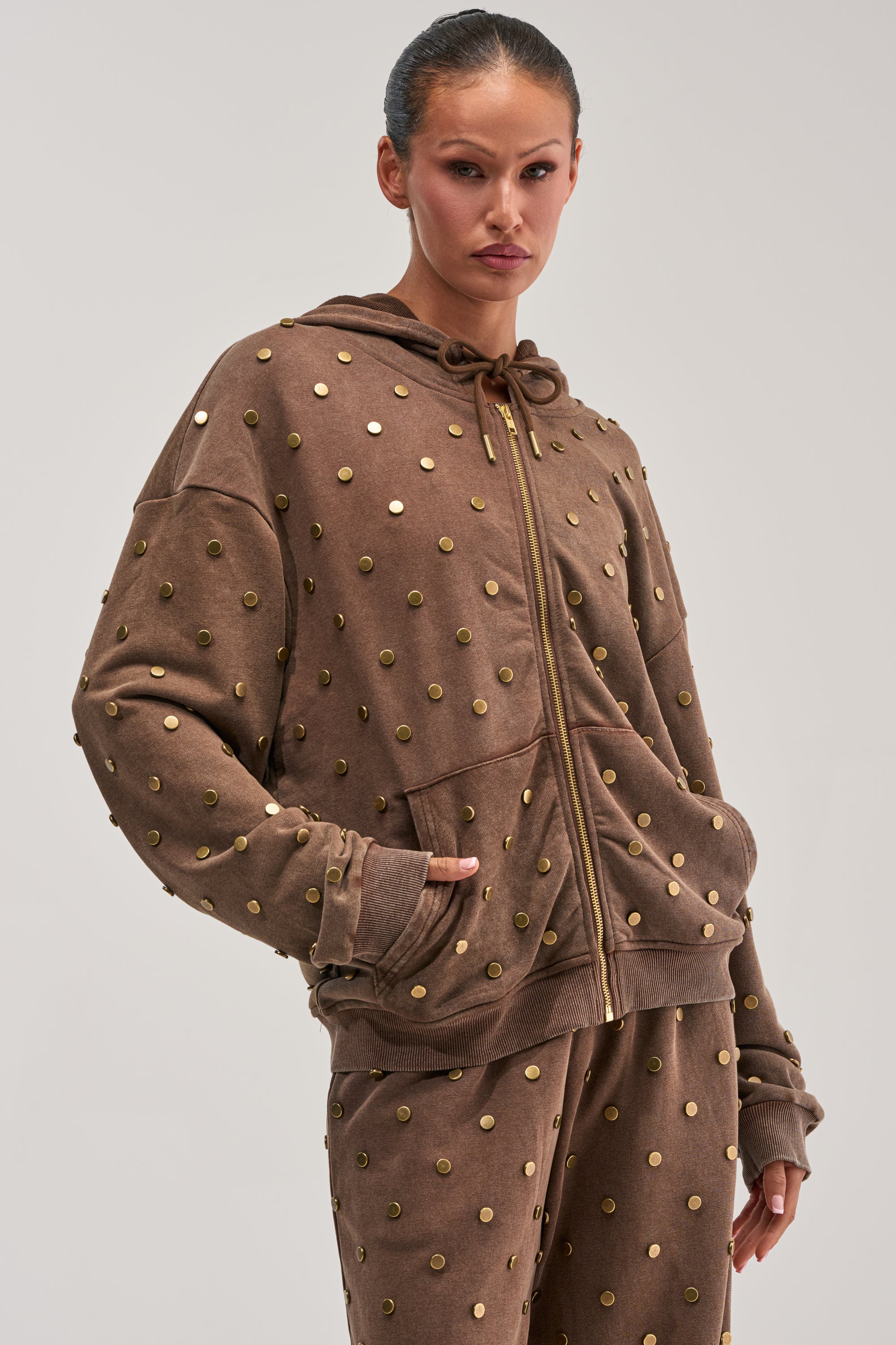 Wearing the POPPING OFF ZIP UP SWEATSHIRT IN BROWN and coordinating studded pants, a woman poses with her hands in her pockets against a plain light background—a striking example of streetwear loungewear.