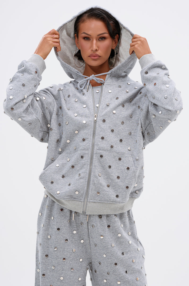 Woman wearing the POPPING OFF ZIP UP SWEATSHIRT with metallic stud embellishments and matching pants, holding the hood with both hands, standing against a plain light background.