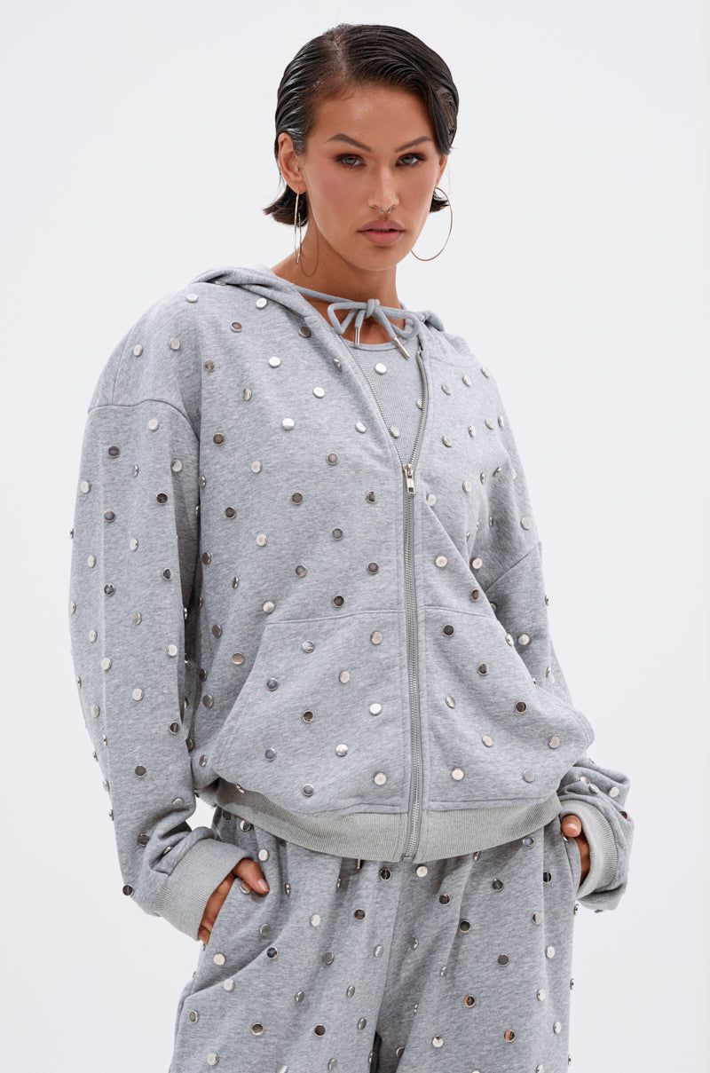 Wearing the POPPING OFF ZIP UP SWEATSHIRT and matching studded sweatpants, a person stands with hands in pockets, confidently facing the camera against a plain white background. Both pieces feature metallic silver stud embellishments.