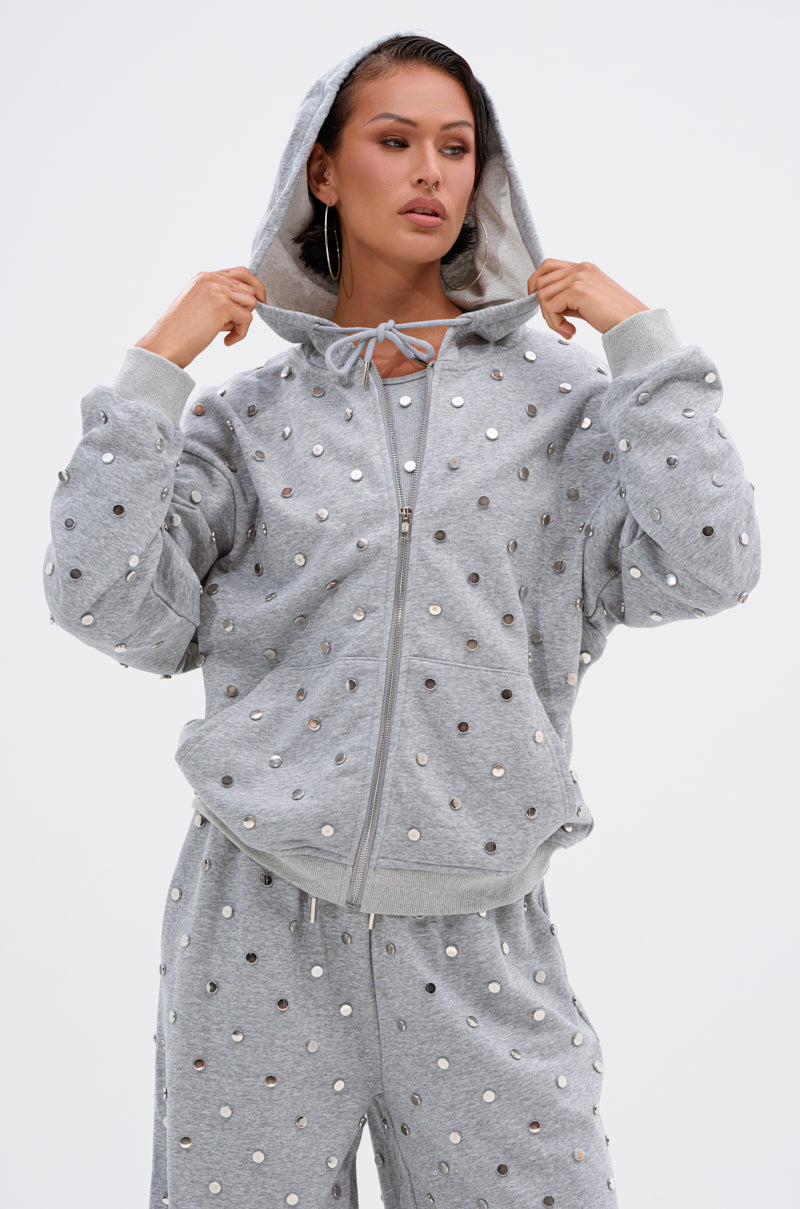 A woman poses against a plain background wearing the POPPING OFF ZIP UP SWEATSHIRT and matching sweatpants, both featuring shiny silver studs, with the hood up and hands gripping the drawstrings.