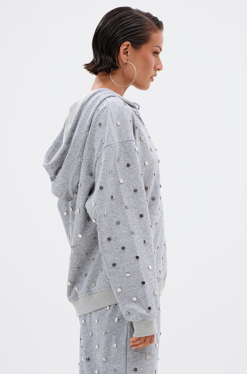 A person with short, slicked-back hair and hoop earrings models the POPPING OFF ZIP UP SWEATSHIRT—a gray, stud-embellished hoodie—paired with matching polka dot pants, shown in profile against a plain white background.