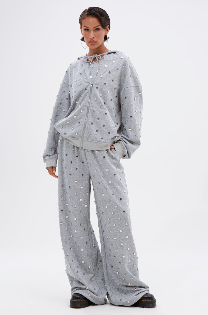 A person stands against a plain background wearing the POPPING OFF ZIP UP SWEATSHIRT, a loose-fitting, light gray hooded tracksuit embellished with shiny silver studs, paired with chunky black shoes.
