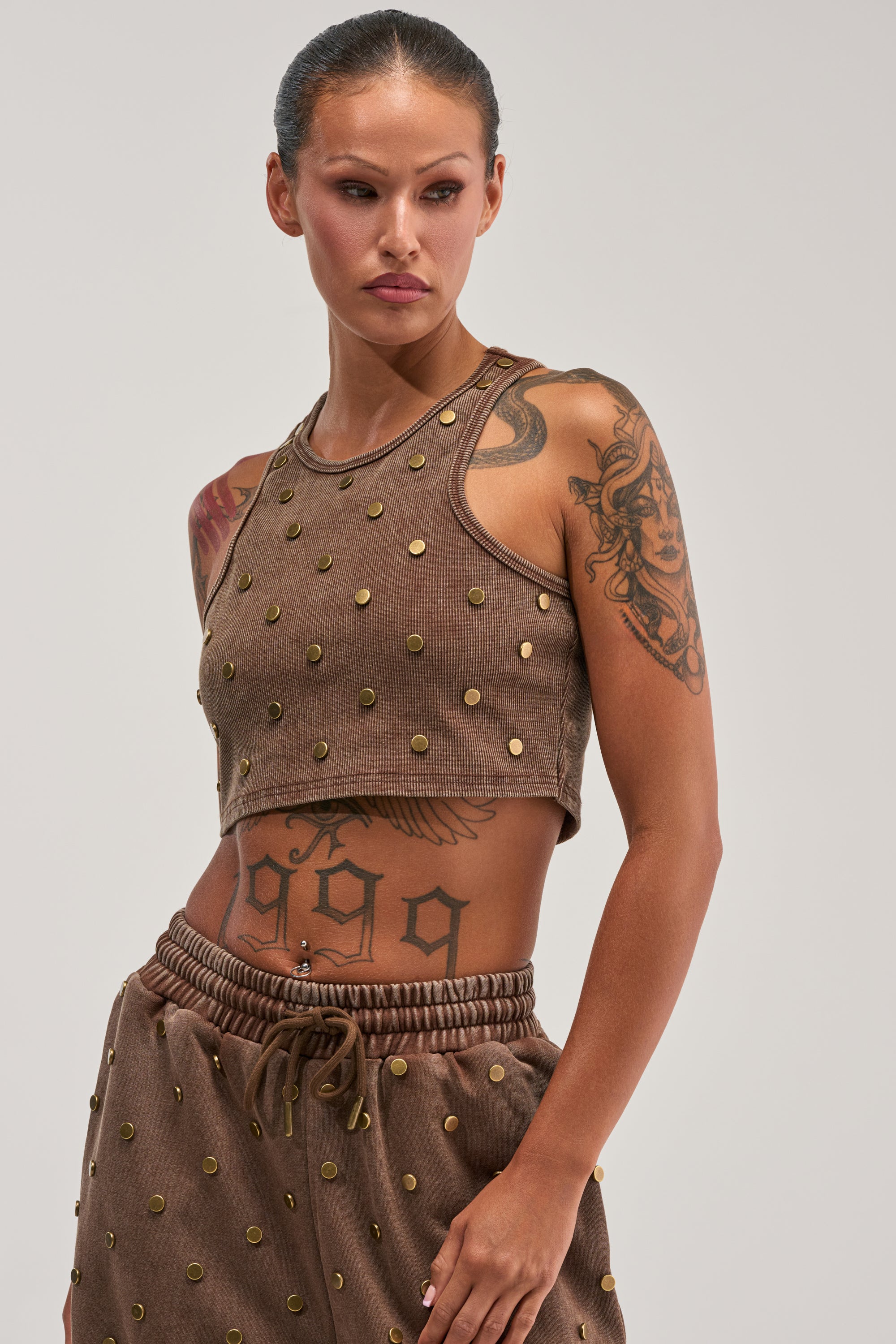 A woman with tattoos wears the POPPING OFF TANK IN BROWN, a ribbed brown crop top with gold stud detailing for bold streetwear style. She pairs it with matching pants and looks to the side, showcasing her tattoo designs against a plain background.