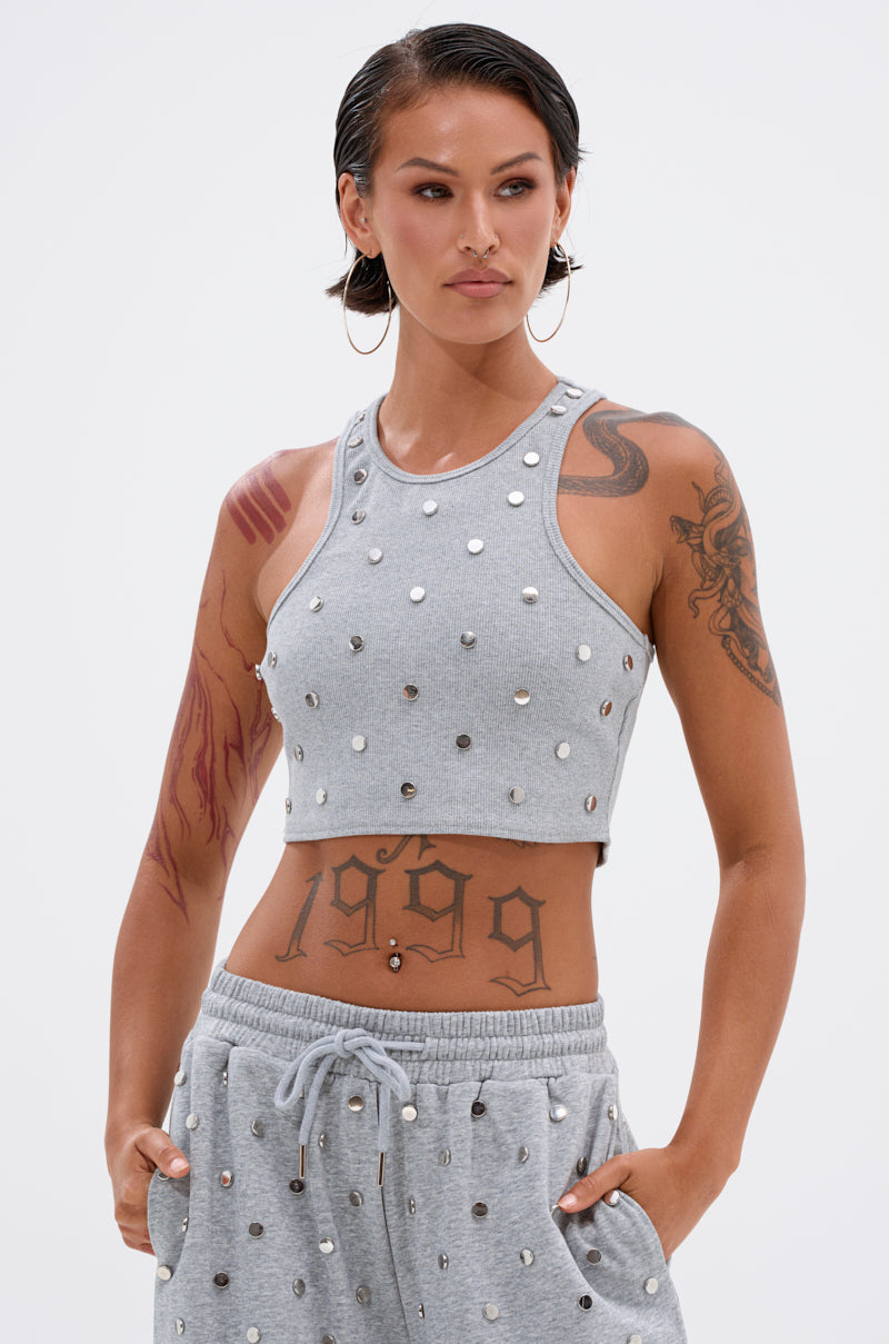 A person with short dark hair and tattoos wears the POPPING OFF TANK and matching sweatpants, both adorned with silver studs. They stand confidently with one hand in their pocket, gazing to the side.