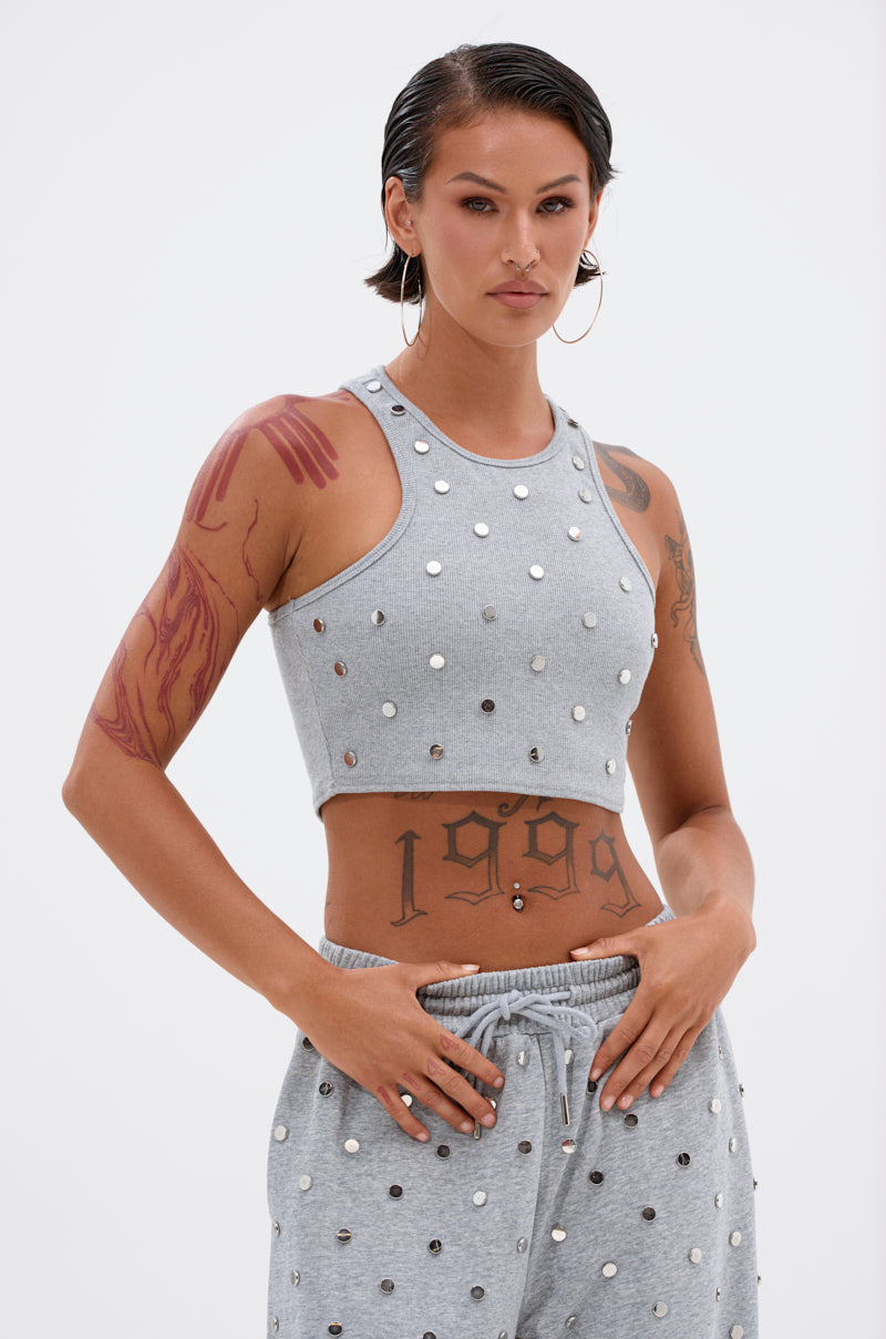 Someone with slicked-back short hair models the POPPING OFF TANK—a grey, stud-embellished crop top—paired with matching studded pants, large hoop earrings, and visible tattoos, standing before a plain light background.