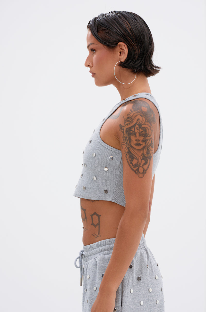 A person with short dark hair, large hoop earrings, and visible tattoos wears the POPPING OFF TANK— a gray stud-embellished top— with matching studded shorts, standing in profile against a plain white background.