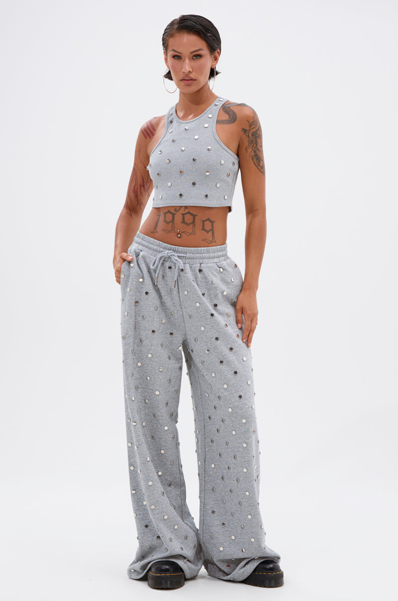 A woman stands confidently in the POPPING OFF TANK, featuring silver studs, with wide-leg sweatpants to match. She displays tattoos on her arms and midriff, accessorized with large hoop earrings and chunky black shoes.