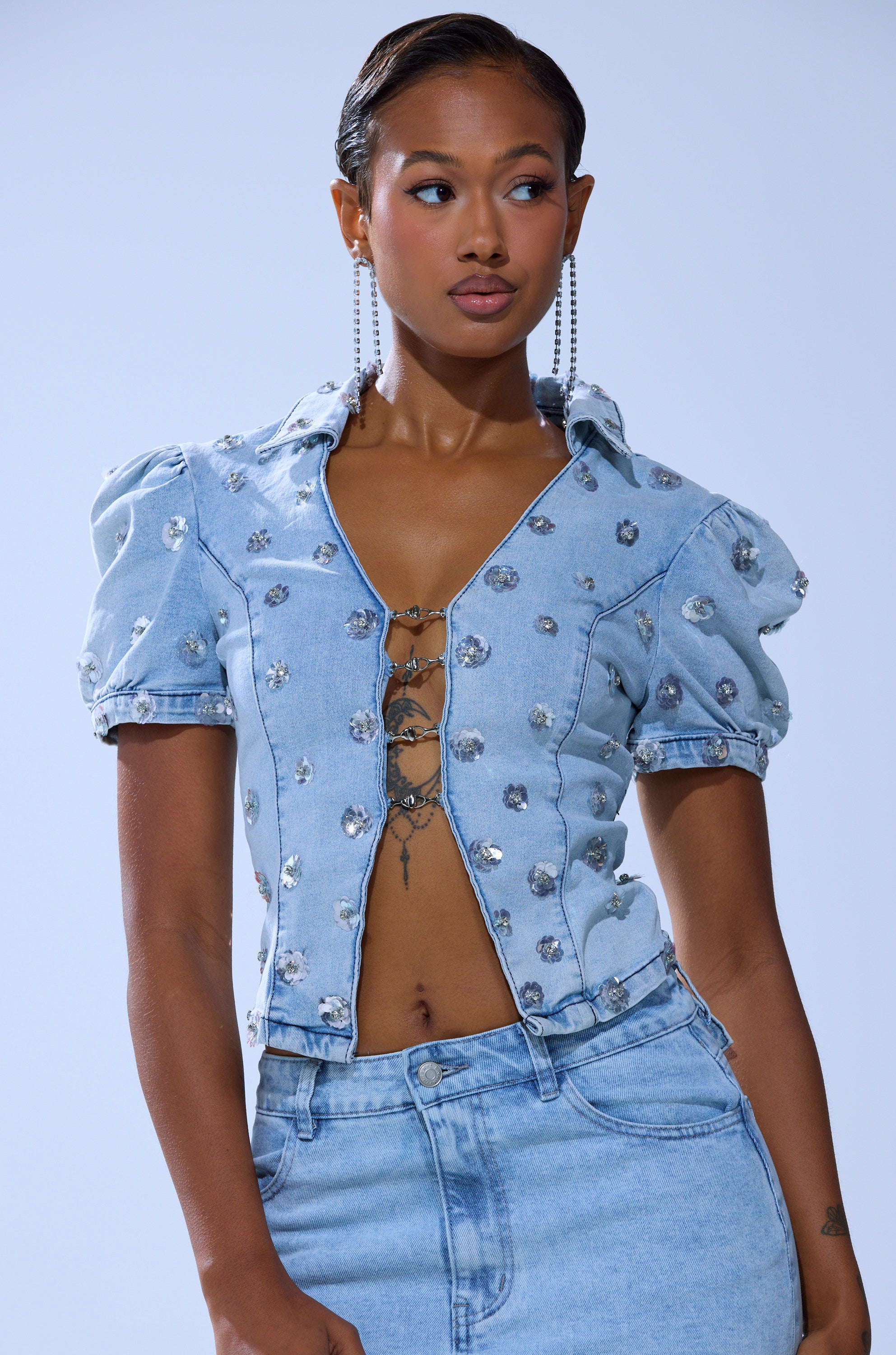 A woman wears the HALEY EMBELLISHED SEQUIN BLOUSE—light blue, short-sleeved with puffed sleeves, floral sequin details, and chain closure—paired with high-waisted denim jeans against a light background.