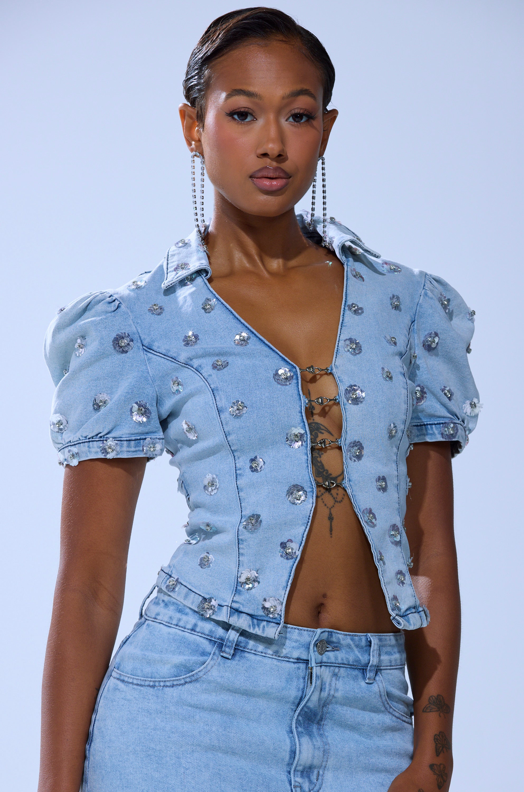 A person wears the HALEY EMBELLISHED SEQUIN BLOUSE—an embellished denim blouse with puffed sleeves, silver details, and front clasps—paired with high-waisted jeans, posing against a plain light background.