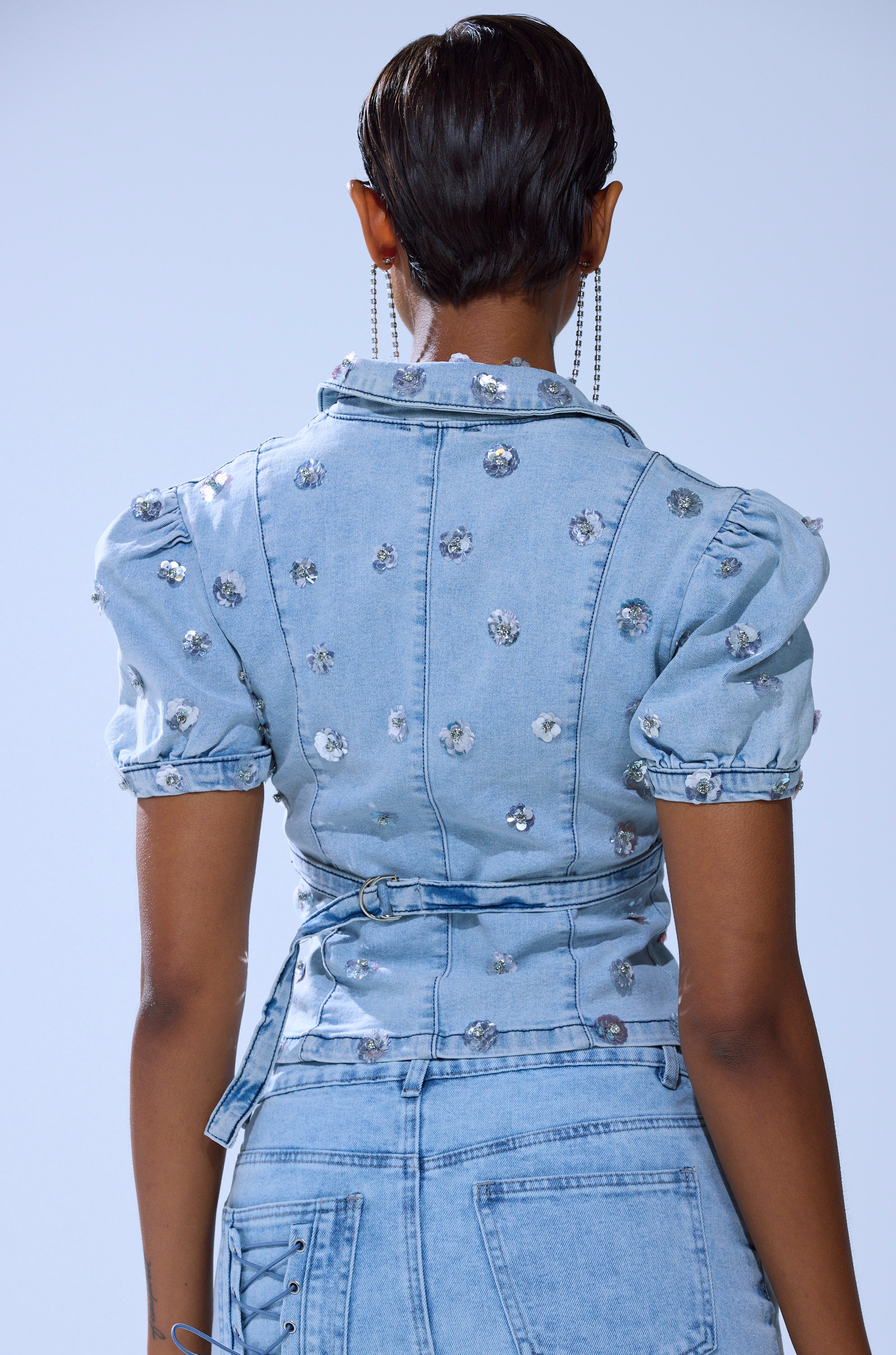 A person with short dark hair, seen from behind, wears the HALEY EMBELLISHED SEQUIN BLOUSE featuring silver details and puffed sleeves, paired with long dangling earrings.