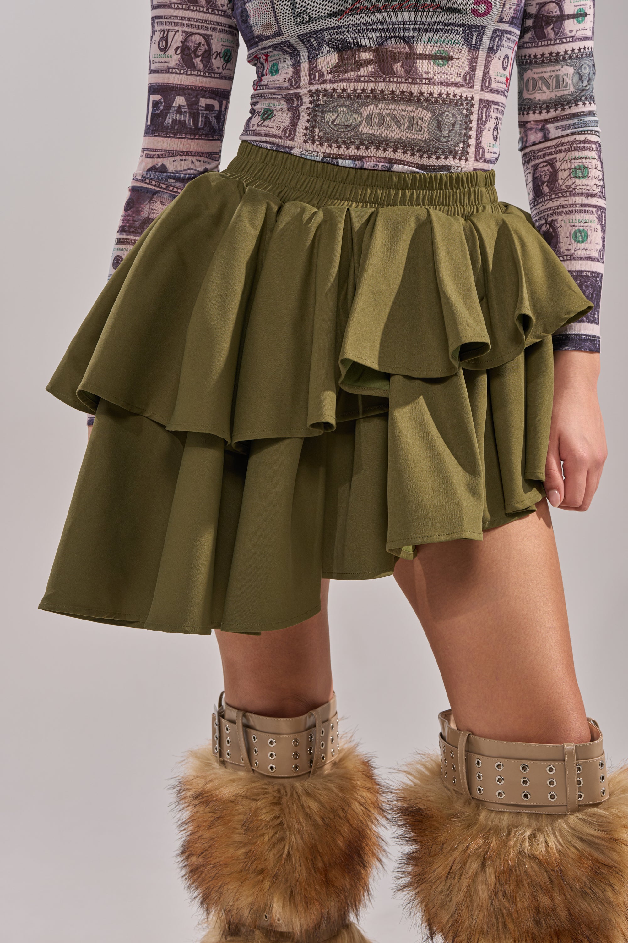 Wearing the OLIVIA OLIVE MINI PLEATED SKIRT with a long-sleeve top featuring a money print and tan fur-trimmed, buckled boots; the photo is cropped from shoulders down. 0