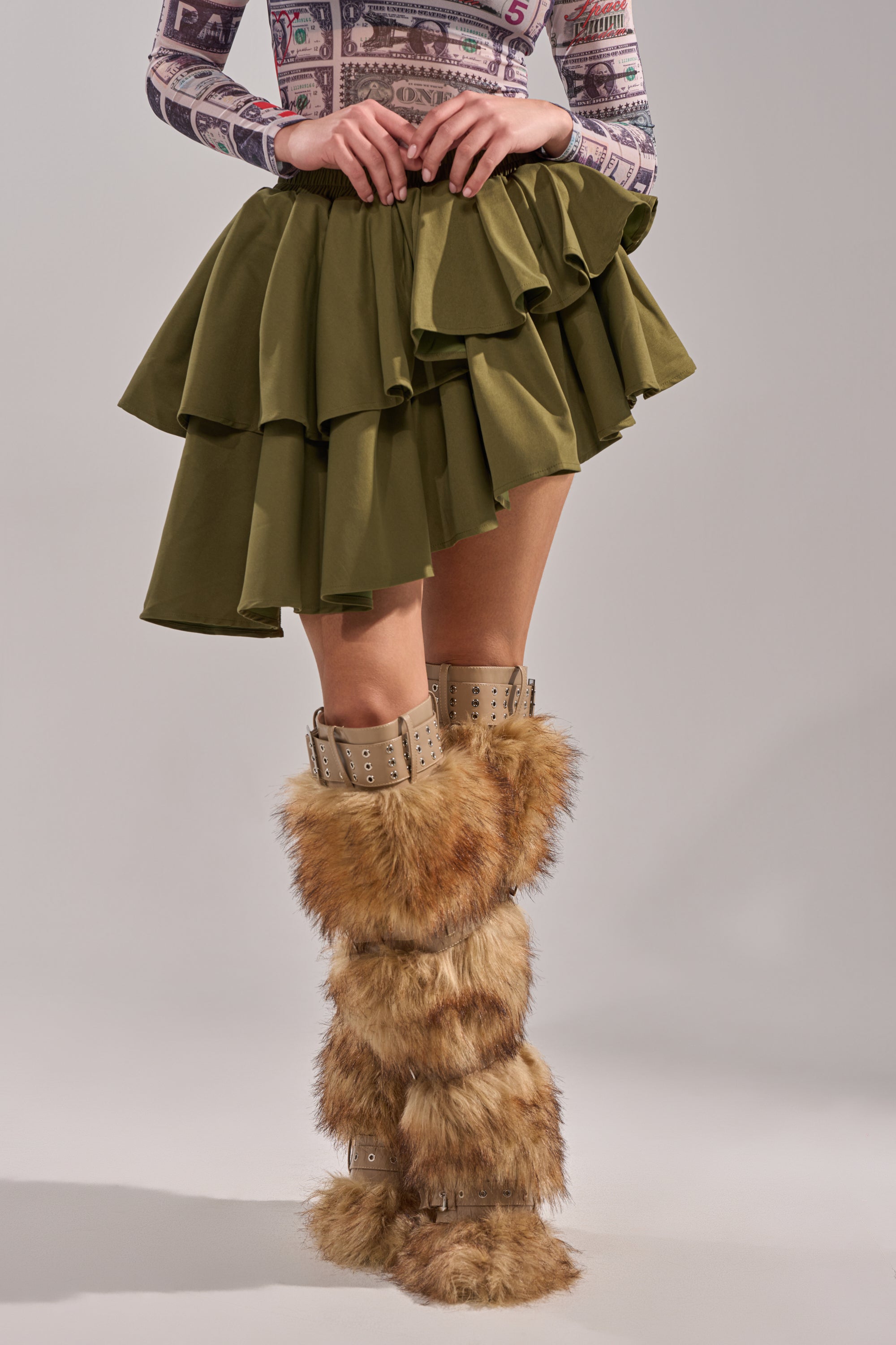 Wearing the OLIVIA OLIVE MINI PLEATED SKIRT with an asymmetric hem, a printed long-sleeve top, and knee-high brown furry boots with buckled straps, a person stands against a plain gray background. 1