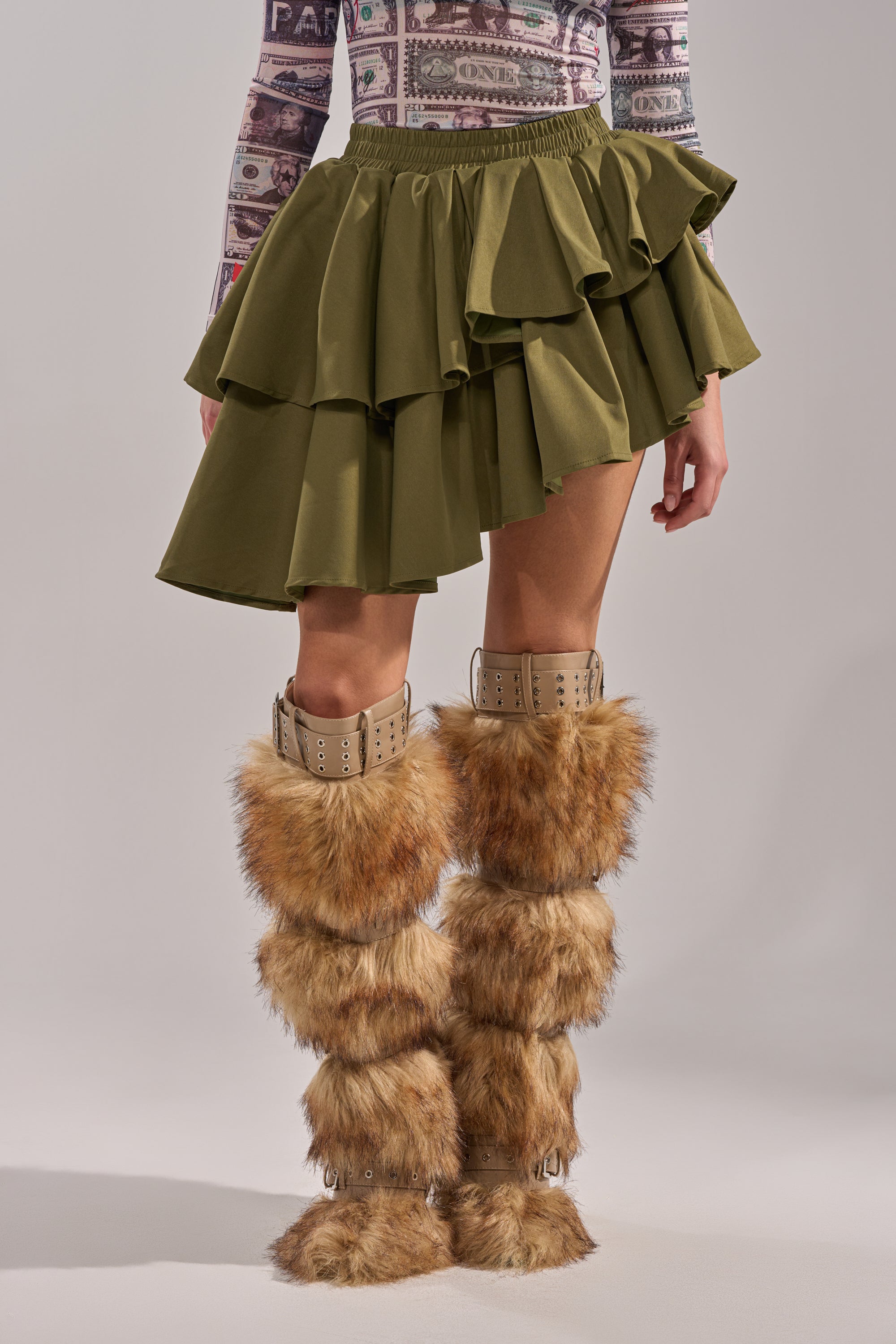 A person wears a patterned long-sleeve top with the OLIVIA OLIVE MINI PLEATED SKIRT, which has an asymmetric hemline, styled with tall tan boots layered with brown faux fur. The image is cropped at the shoulders. 2