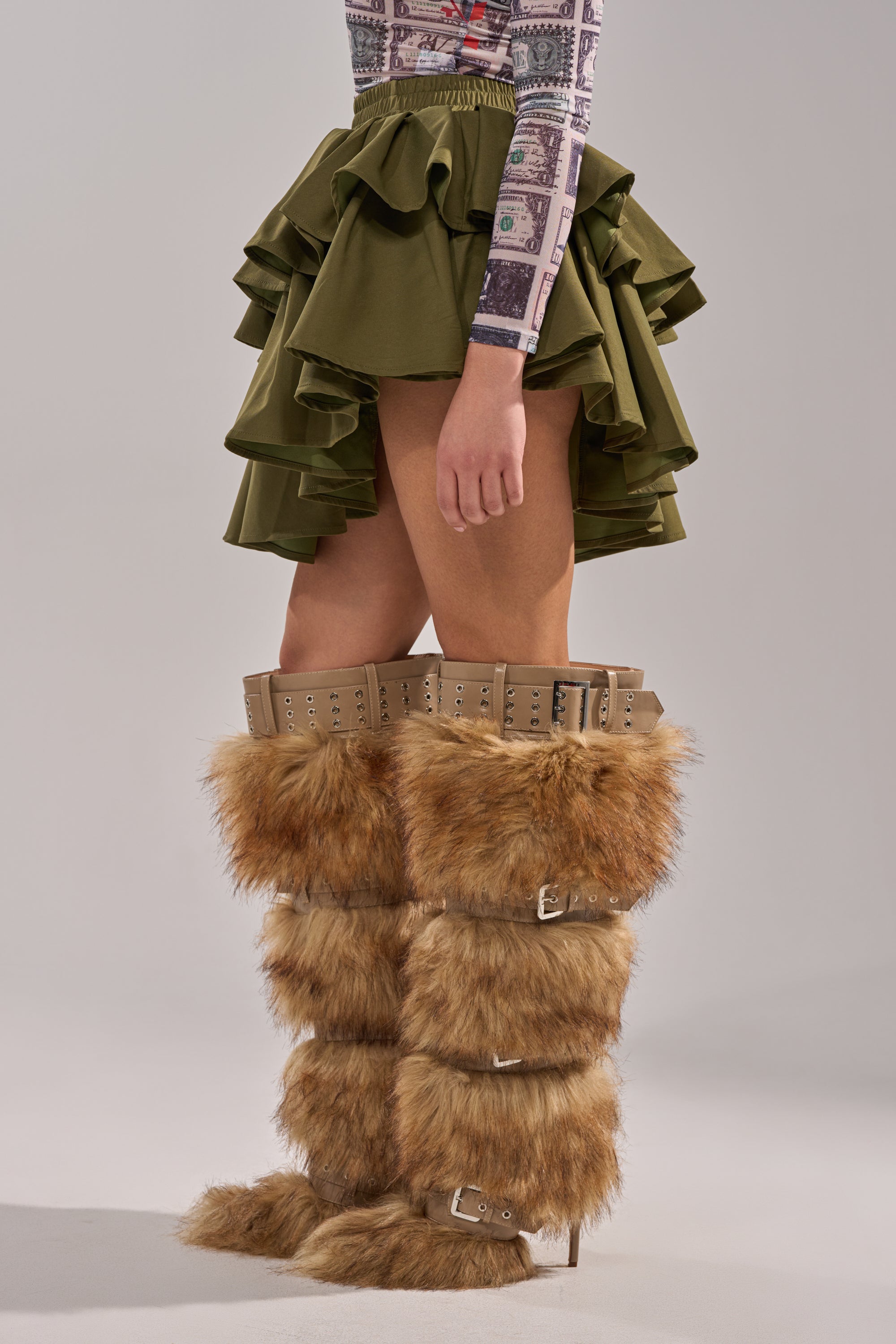 Wearing the OLIVIA OLIVE MINI PLEATED SKIRT with layered pleats and tall tan faux fur boots with multiple buckles, a person stands against a plain light background, showing only their lower half. 3