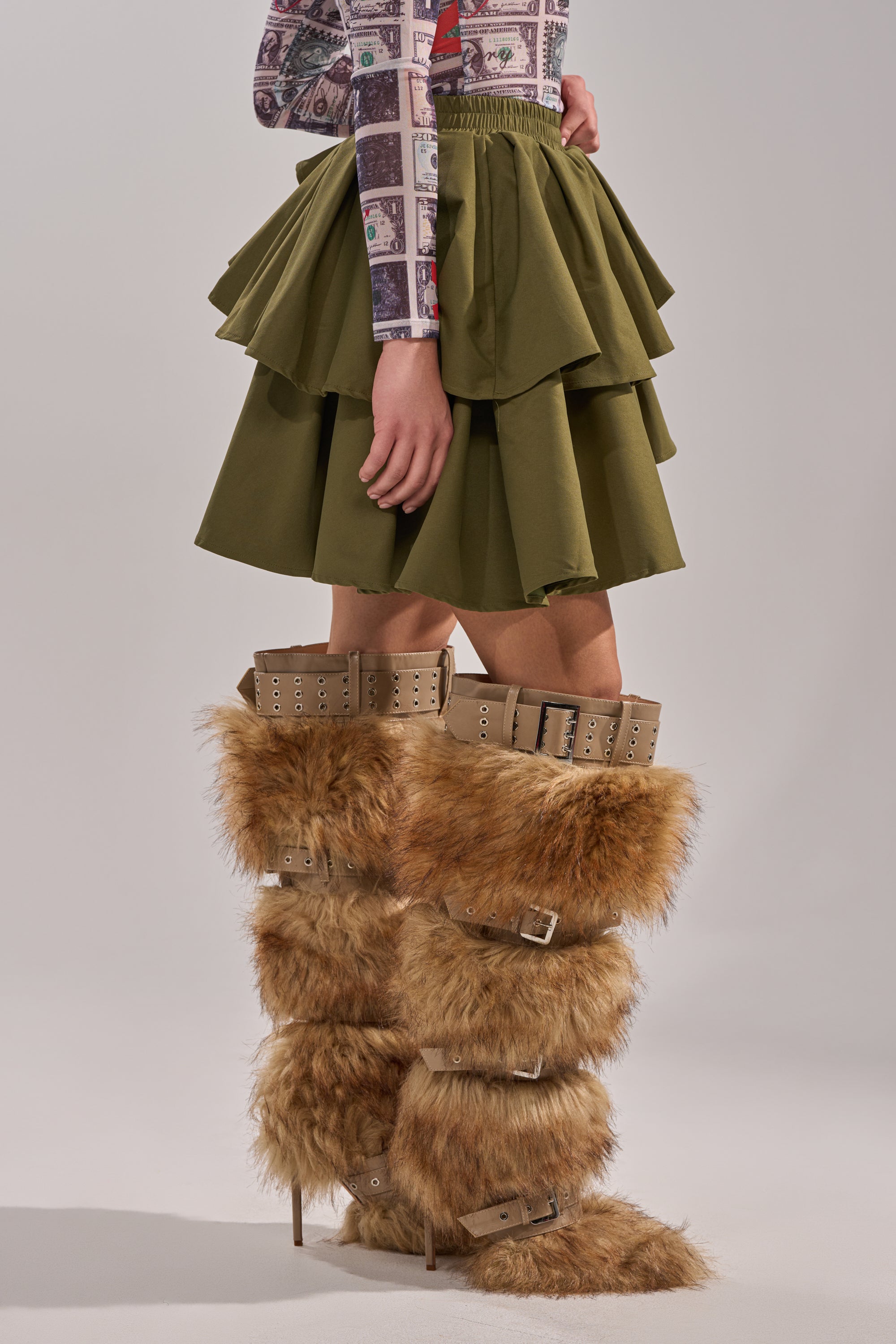 A person stands against a plain background wearing the OLIVIA OLIVE MINI PLEATED SKIRT with layered pleats, a long-sleeve printed top, and tall tan boots featuring shaggy faux fur and buckled straps. 4