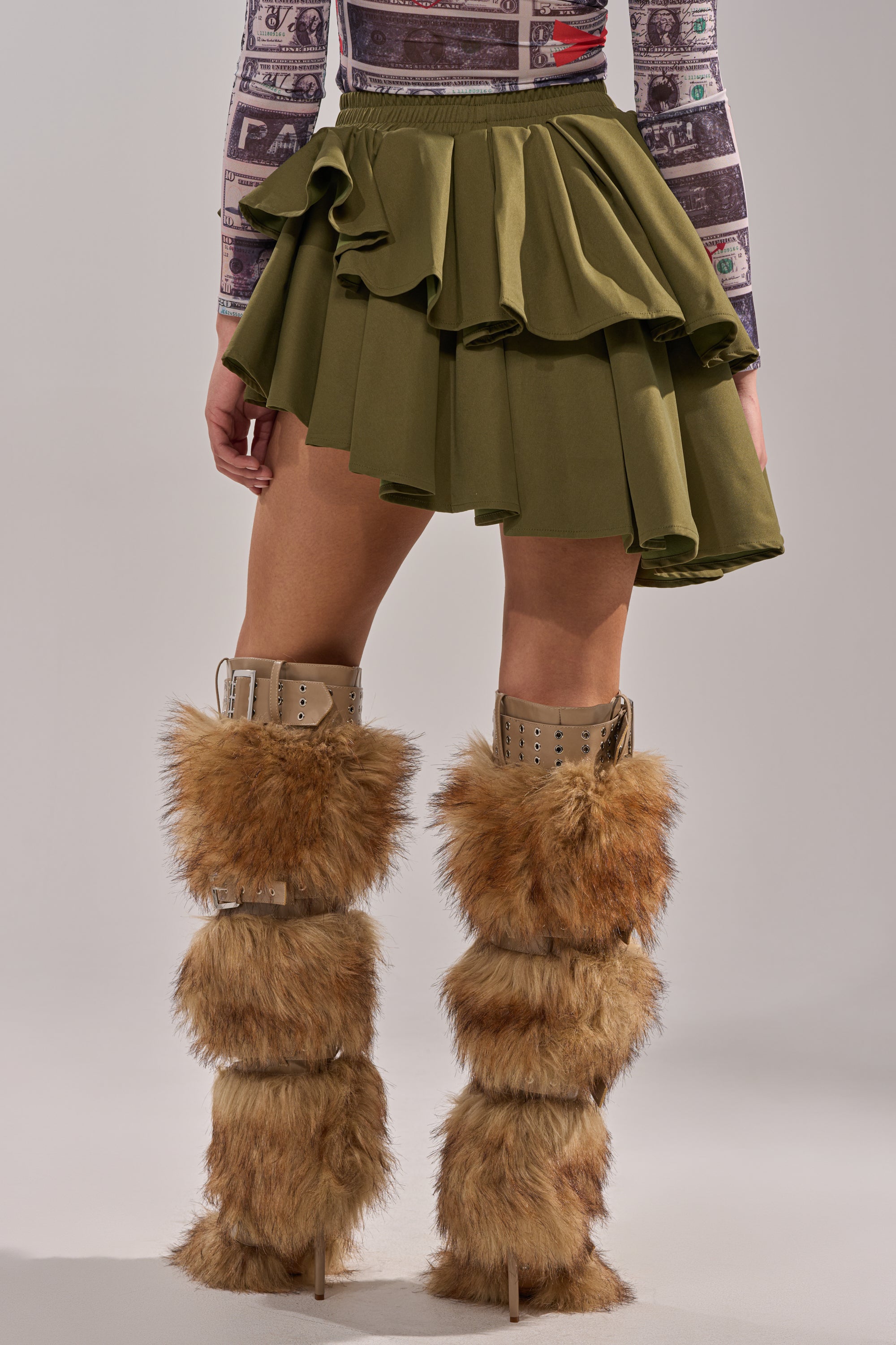 A person stands against a plain background wearing the OLIVIA OLIVE MINI PLEATED SKIRT with an asymmetric hem, layered brown faux fur knee-high boots, and a patterned top featuring various images and text. 5
