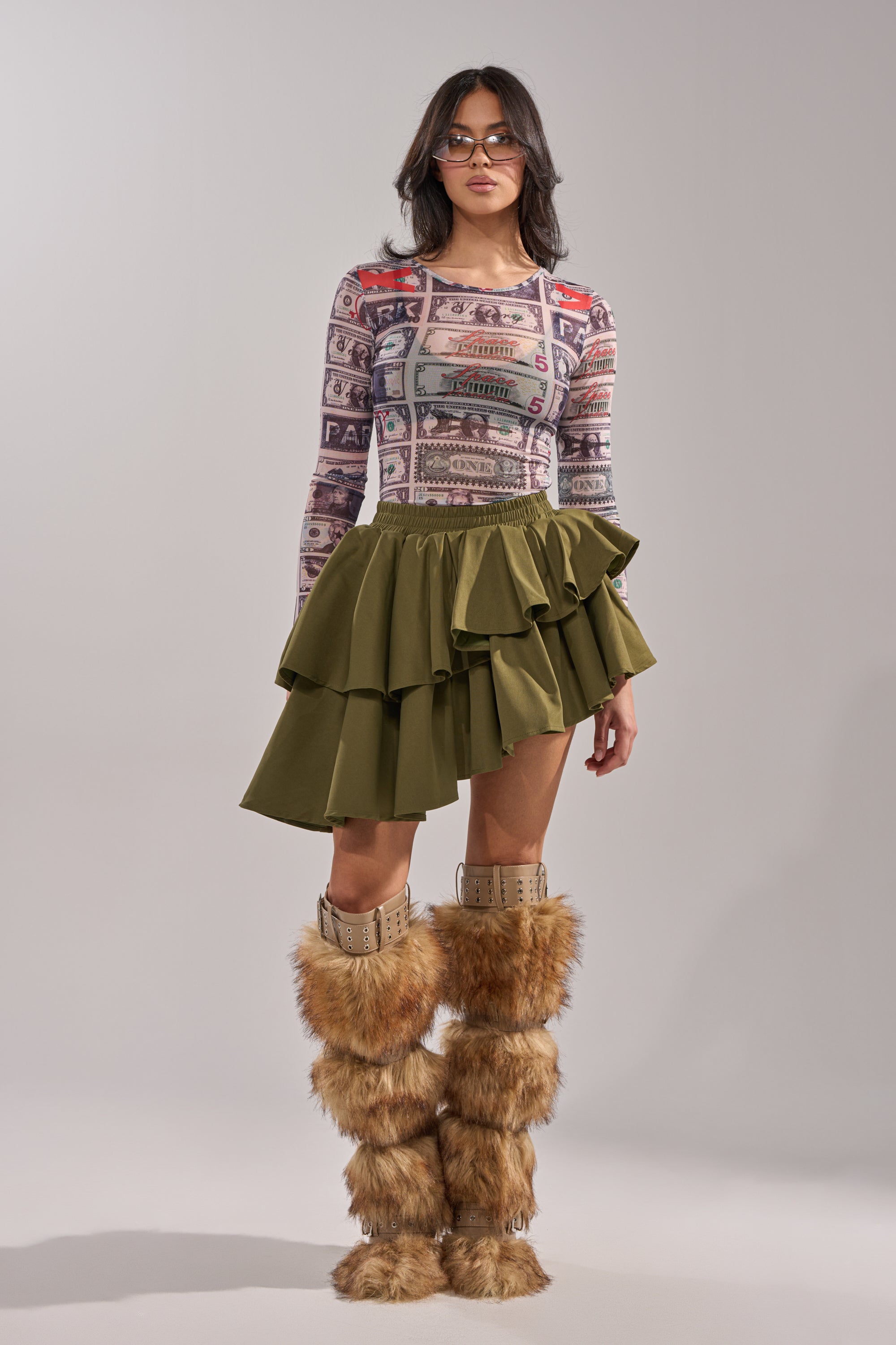 A woman in sunglasses, a colorful graphic long-sleeve top, the OLIVIA OLIVE MINI PLEATED SKIRT, and tall furry brown boots stands against a plain gray background. 6
