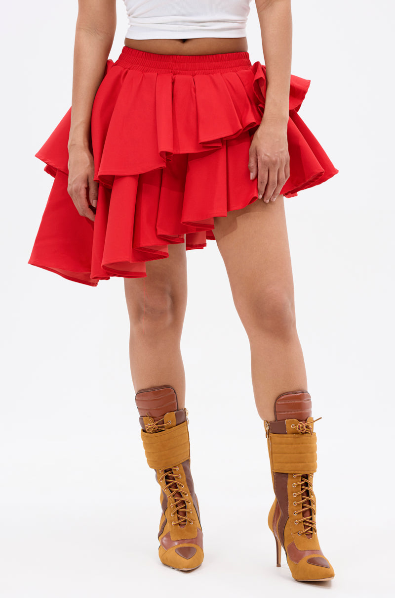 A person models the RUBY RED MINI PLEATED SKIRT with layered pleats, paired with a white top and tan lace-up high-heeled boots, standing against a plain white background. Their face is not visible.