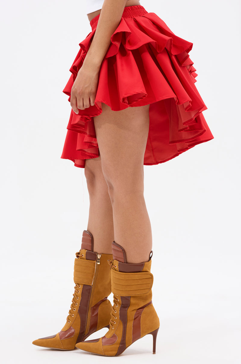 A person stands sideways wearing the RUBY RED MINI PLEATED SKIRT with tan lace-up heeled boots with brown accents, set against a plain white background.