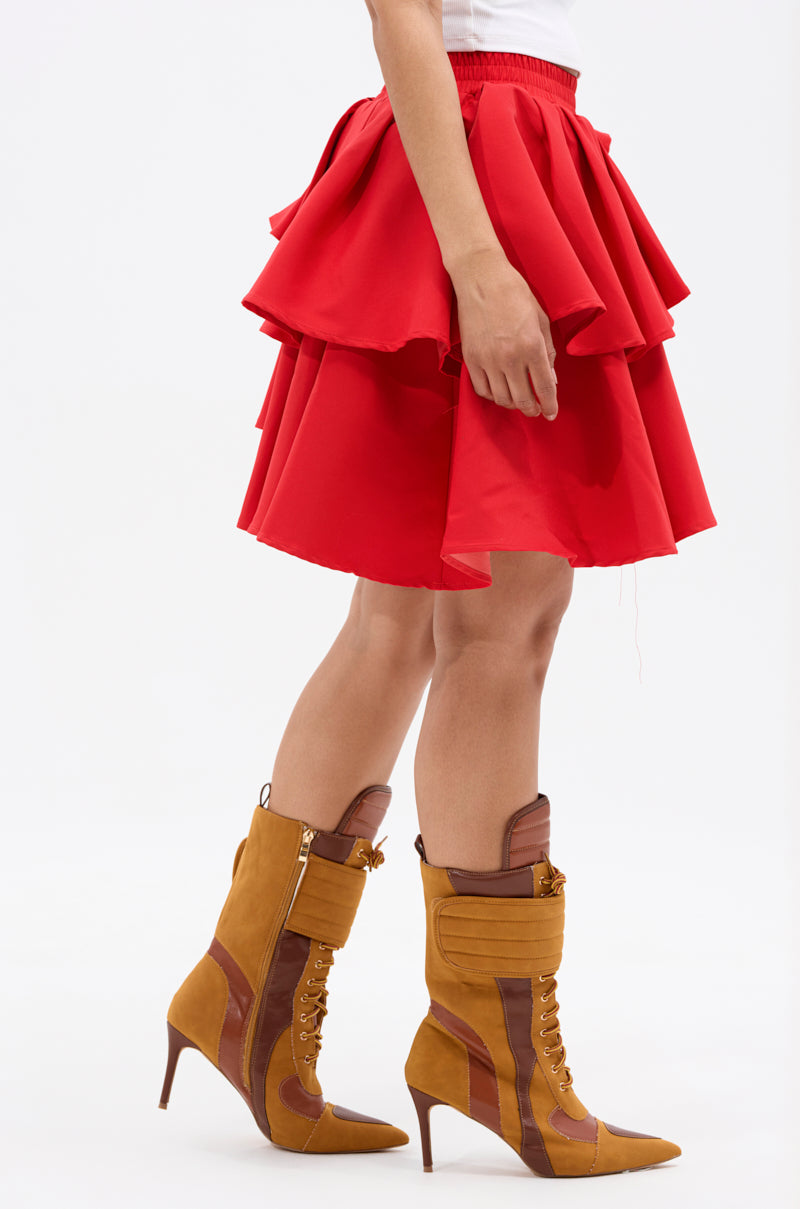 Model shown from the waist down wears the RUBY RED MINI PLEATED SKIRT with layered pleats, styled with high-heeled lace-up brown and mustard boots against a plain white background.