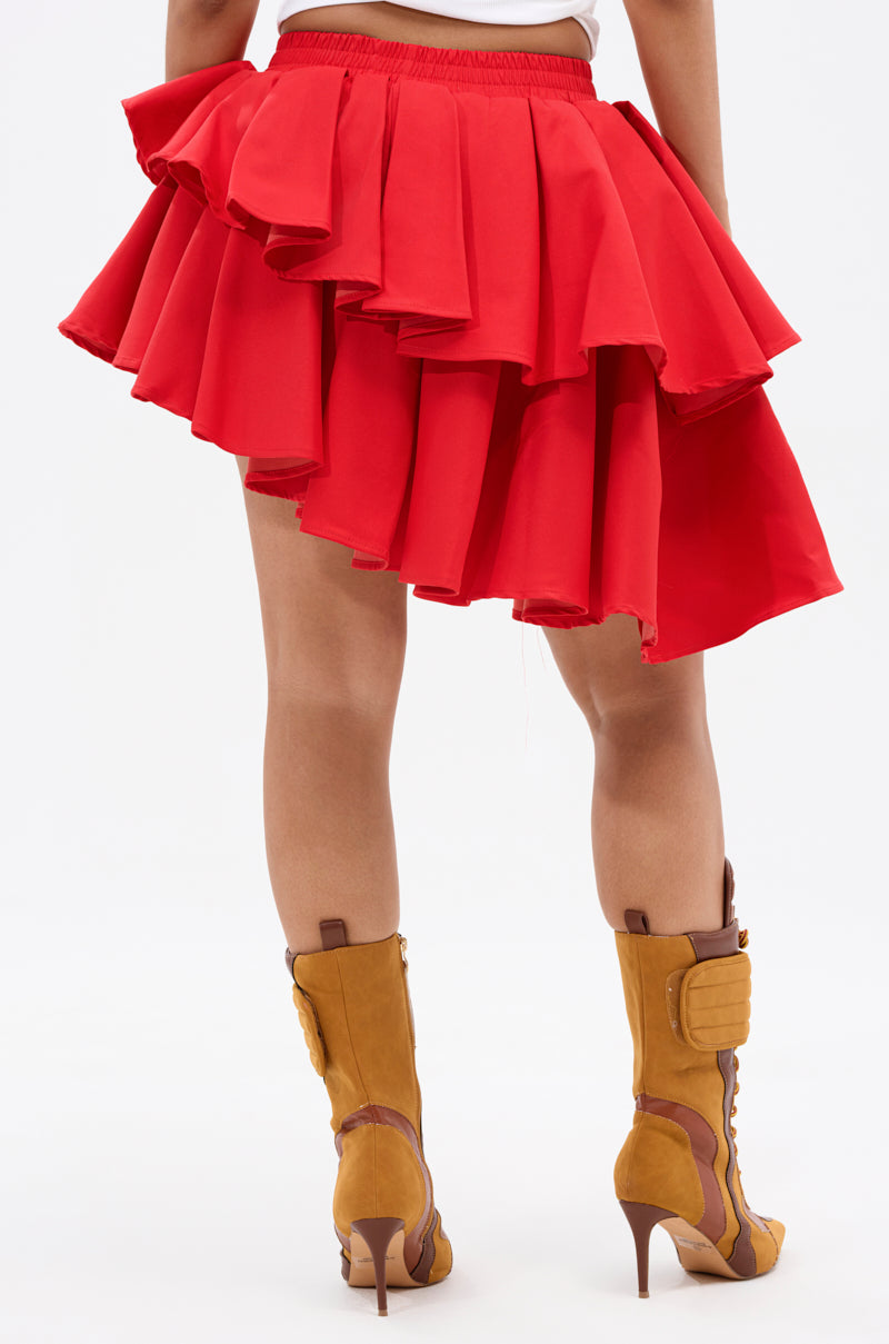 A person is seen from the back in a RUBY RED MINI PLEATED SKIRT with layered pleats and tan high-heeled boots, standing against a plain white background.