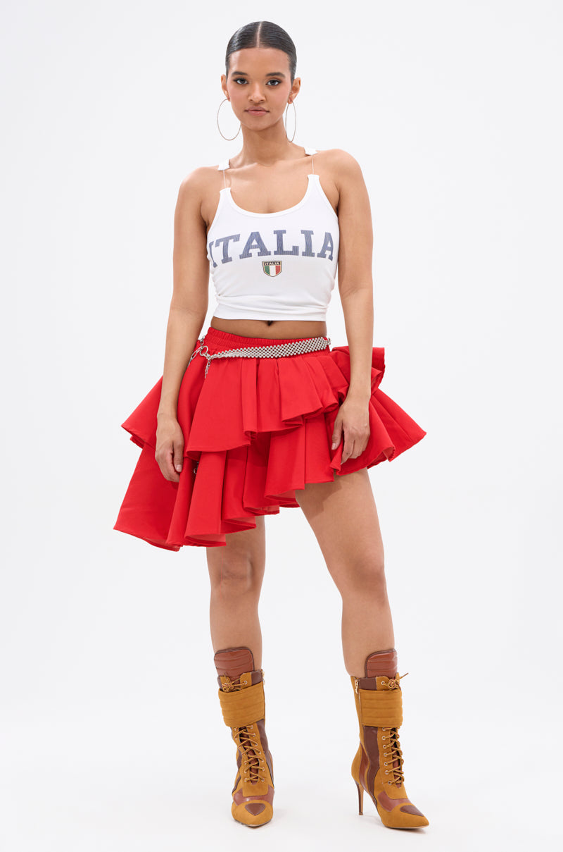 A woman faces forward against a white background, wearing a white "ITALIA" tank top, the RUBY RED MINI PLEATED SKIRT with an elastic waistband, a chain belt, brown lace-up heeled boots, hoop earrings, and her hair pulled back.