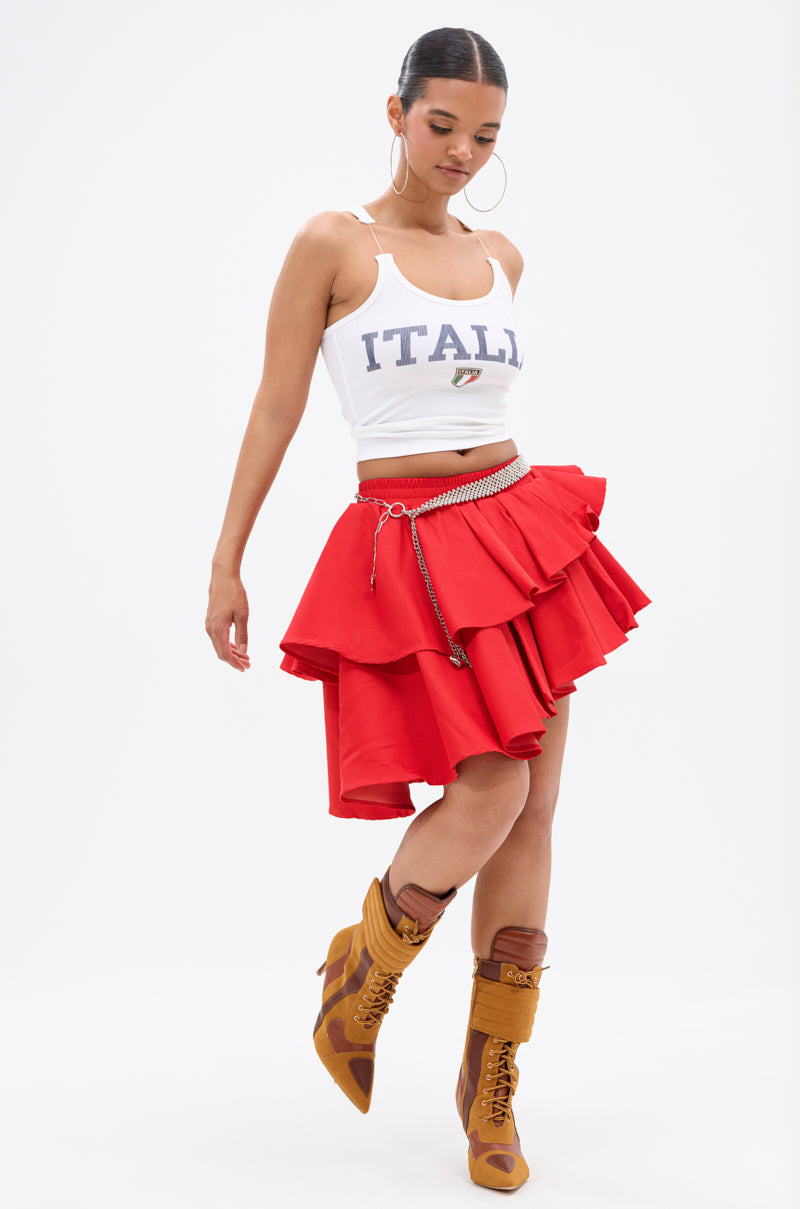 A woman in a white "ITALIA" tank top, RUBY RED MINI PLEATED SKIRT with elastic waistband, chain belt, tan lace-up boots, and large hoop earrings poses against a plain white background.