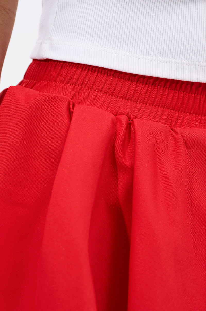 Close-up of the elastic waistband and gathered fabric of the RUBY RED MINI PLEATED SKIRT, featuring a smooth, shiny texture and an asymmetric hemline, paired with a white ribbed top.