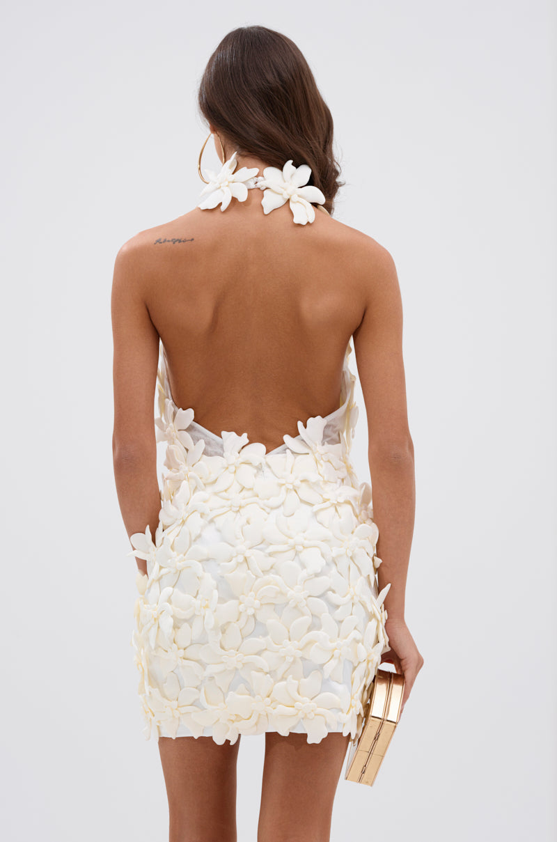 A woman with long brown hair is seen from behind in the ASHLEY FLORAL MINI DRESS IN WHITE, an open-back mini dress with large floral appliqués. She holds a gold clutch and shows off a tattoo on her left shoulder—perfect for garden parties.