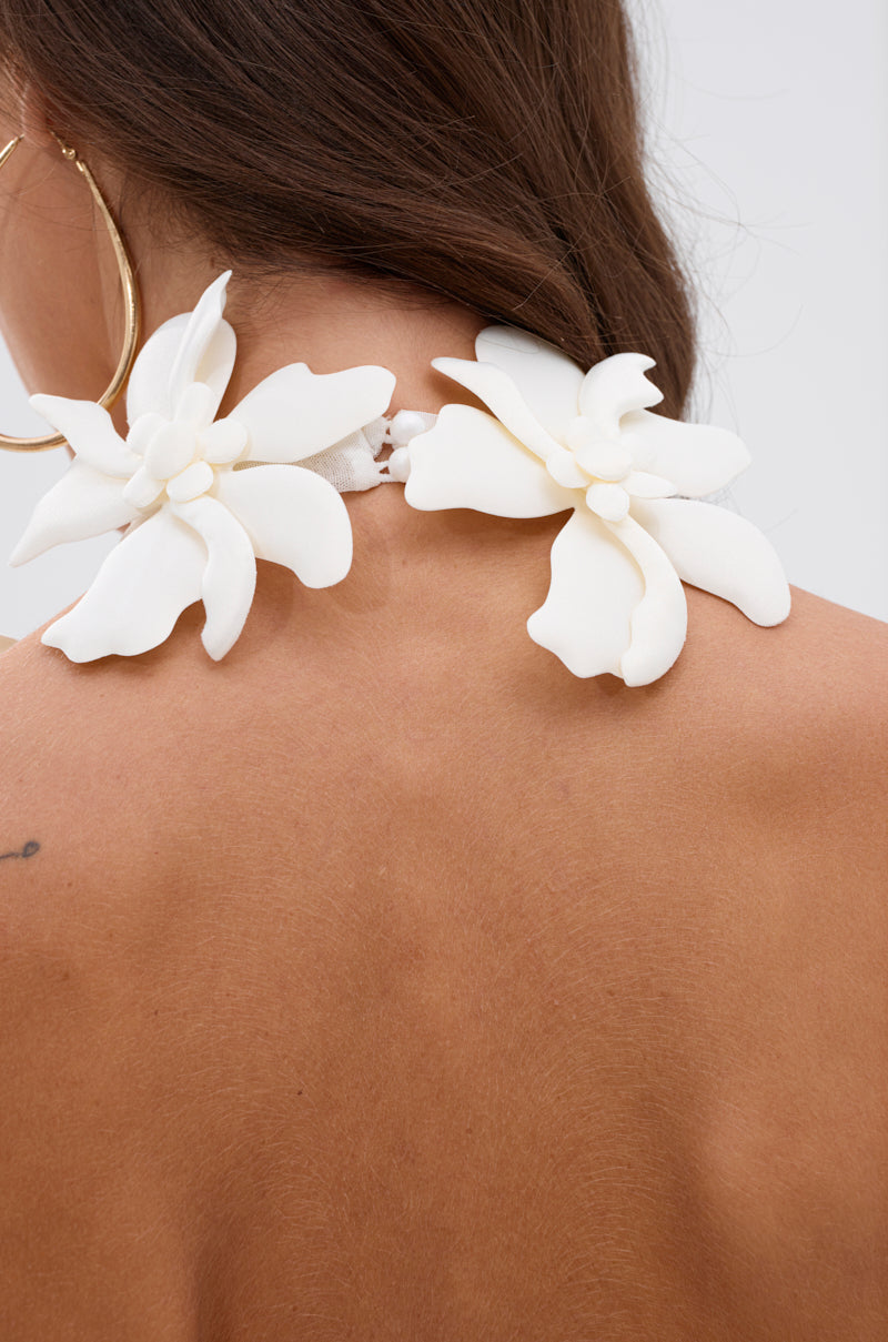 Close-up of a woman's bare back and shoulder with two large white floral accessories near her neck, gold hoop earring, and brown hair draped over one shoulder—a stylish look paired with the ASHLEY FLORAL MINI DRESS IN WHITE.