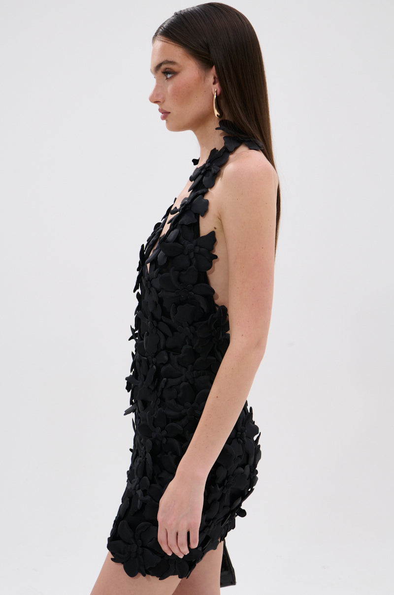 A woman with straight brown hair poses in profile against a white background, wearing the ASHLEY FLORAL MINI DRESS IN BLACK, a sleeveless mini dress featuring 3D floral appliqués.