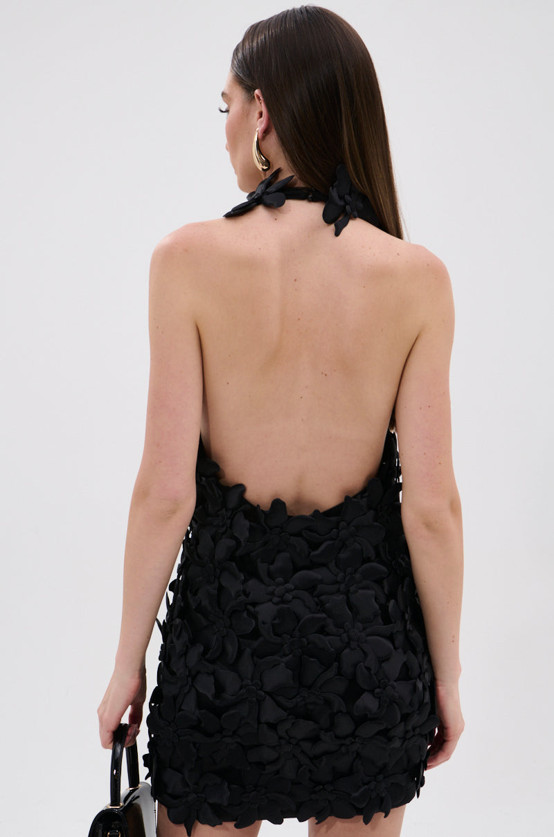 A woman with long, straight brown hair models the ASHLEY FLORAL MINI DRESS IN BLACK, a mesh halter mini dress with 3D floral appliqués and an open back. She holds a black handbag and faces away against a plain background.
