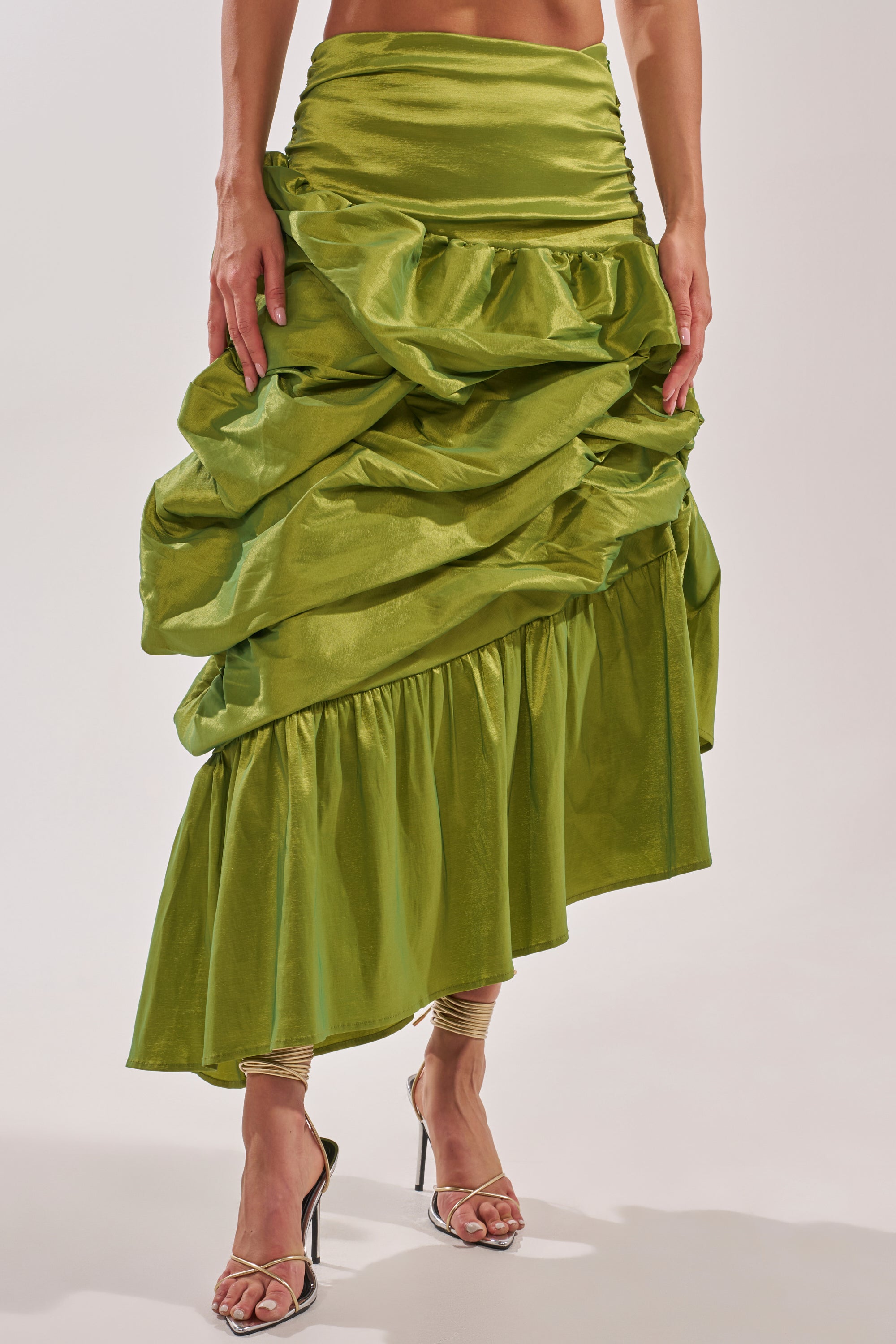A person wearing the ALL OVER THE PLACE MIDI SKIRT—a shiny, bright green ruched midi with ruffled layers and an asymmetrical hem—paired with strappy high-heeled sandals. Only the skirt and lower body are shown. 0