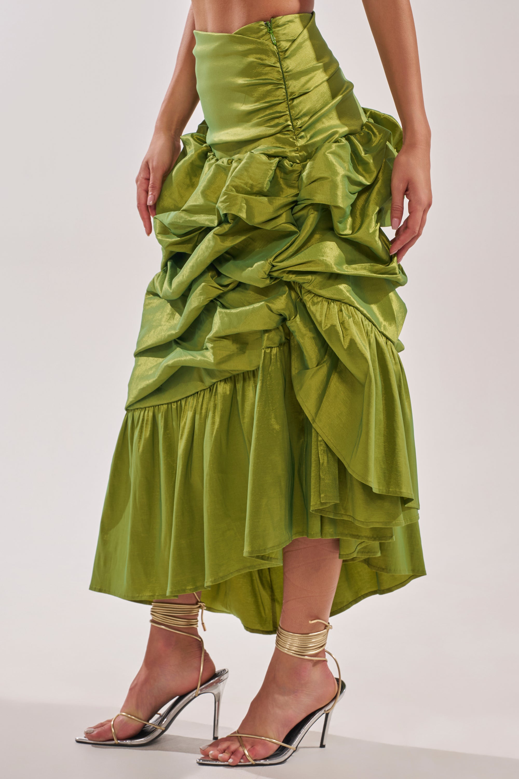 Wearing the ALL OVER THE PLACE MIDI SKIRT—a shiny green ruched skirt with ruffles and an asymmetrical hem—paired with metallic gold heels. The photo is cropped at the midriff, highlighting both the stylish skirt and shoes. 2