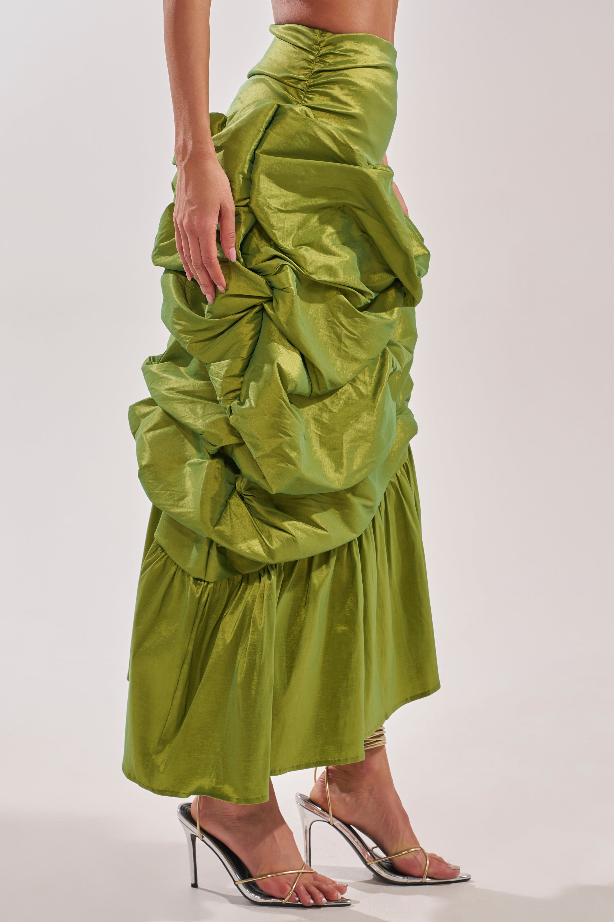 Someone wears the ALL OVER THE PLACE MIDI SKIRT, a shiny green ruched midi with voluminous texture and graceful gathers, paired with metallic high-heeled sandals, standing out against a plain light background. 3