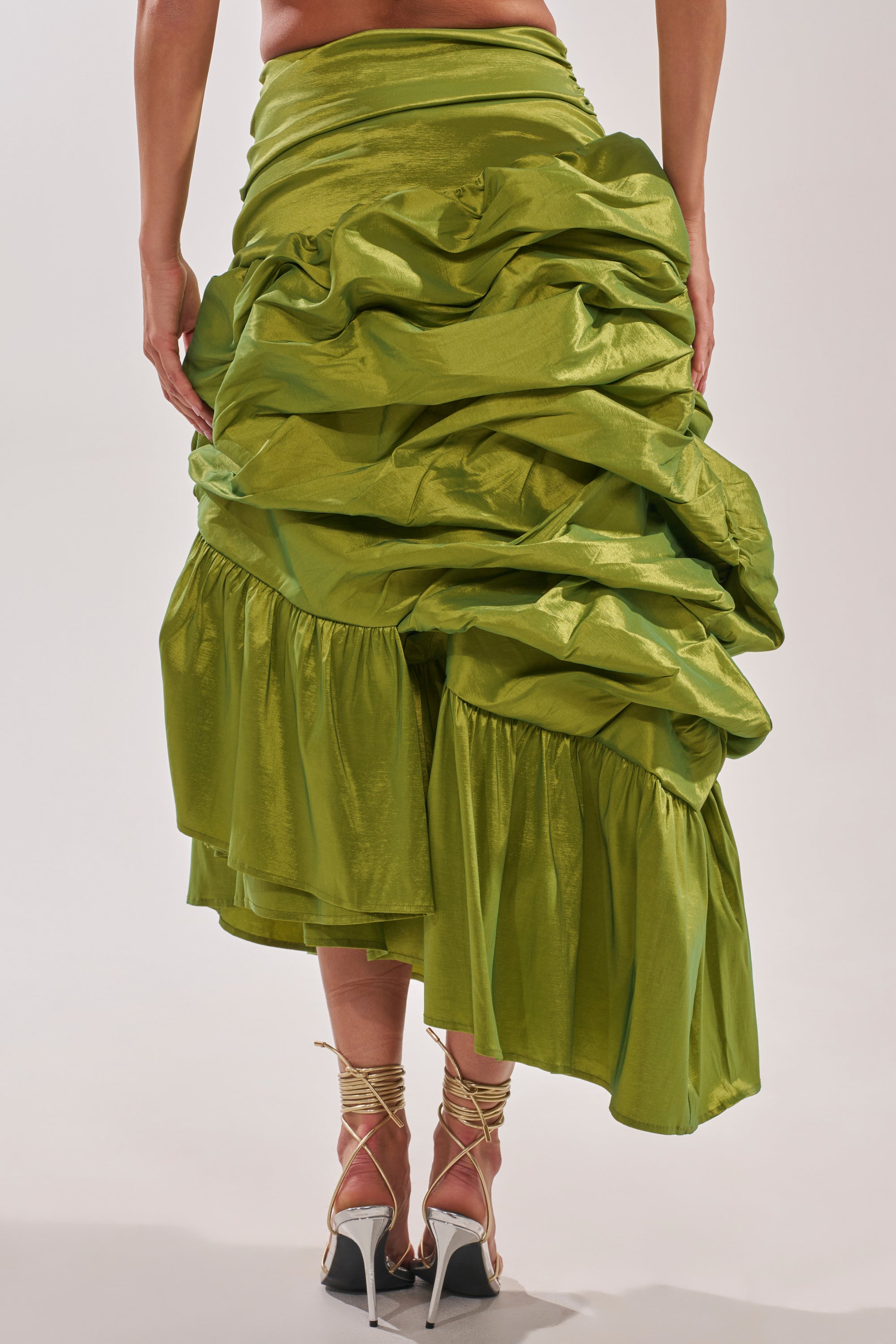 A person wearing the ALL OVER THE PLACE MIDI SKIRT, a shiny green ruched skirt with an asymmetrical hem, paired with strappy high-heeled sandals. The image shows the back view from waist to feet against a plain background. 4