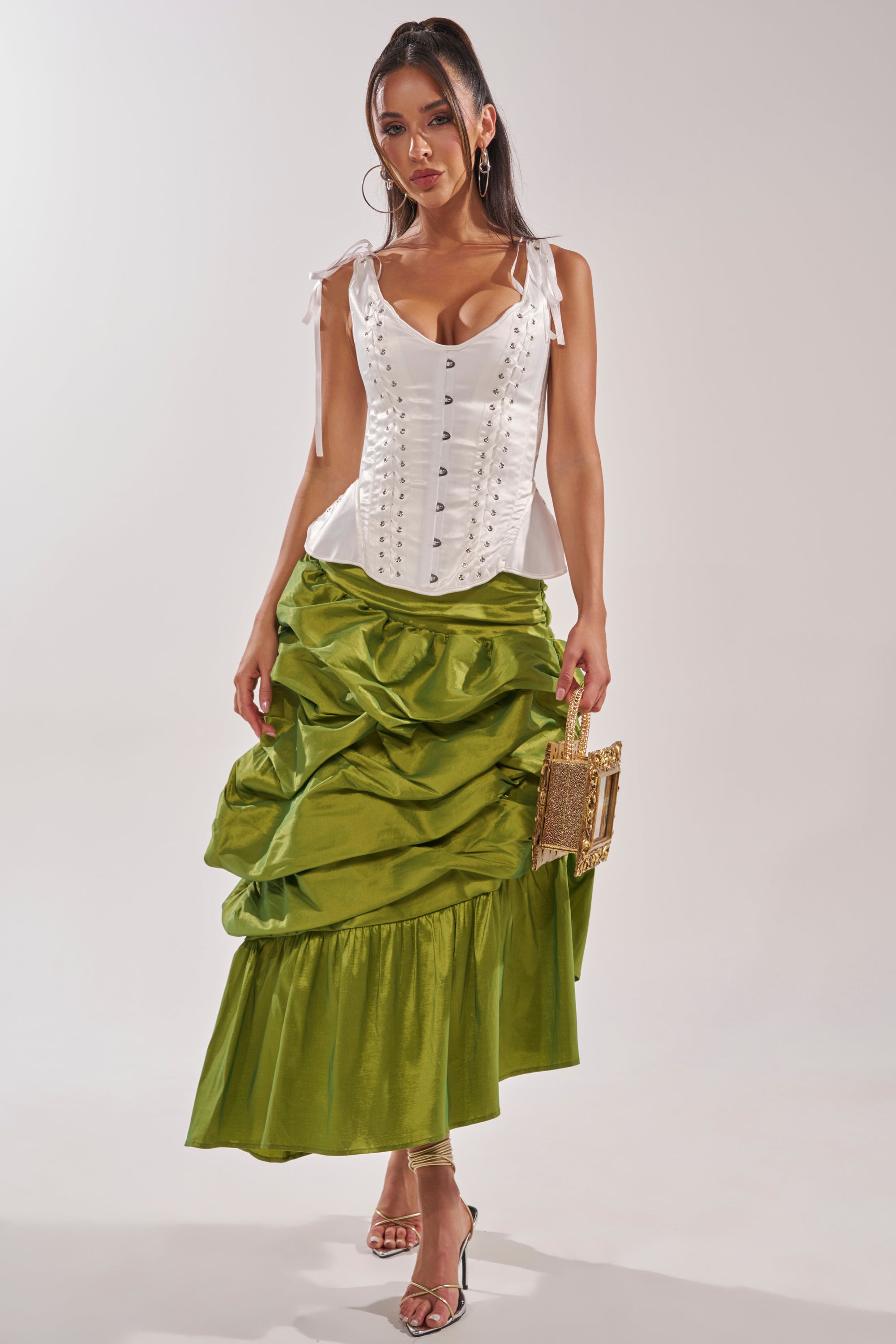 A woman poses against a plain background in the ALL OVER THE PLACE MIDI SKIRT, paired with a white corset top. She holds a square handbag, wears clear high-heeled sandals, and her long straight hair completes the chic look. 5